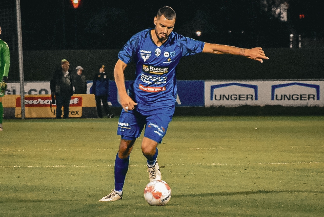 Fußballspieler in blauem Trikot kontrolliert den Ball auf dem Spielfeld bei Nachtspiel.
