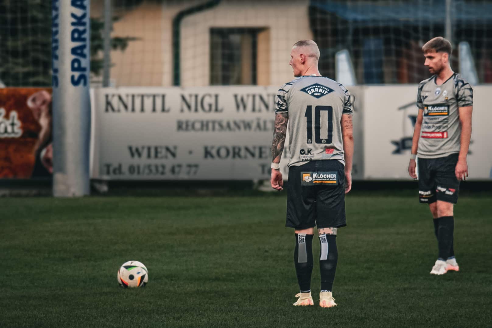 Fußballspieler auf dem Platz, bereit für einen Freistoß während eines Spiels, Beratungen im Hintergrund.