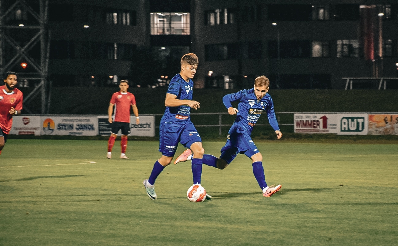 Zwei Fußballspieler im blauen Trikot spielen auf einem beleuchteten Fußballfeld bei Nacht gegeneinander.