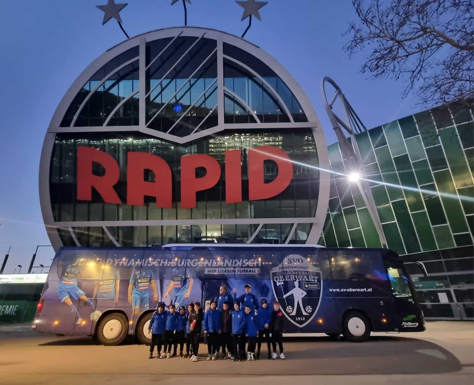 Fußballteam vor dem Rapid-Stadion mit Team-Bus im Vordergrund bei Abenddämmerung.