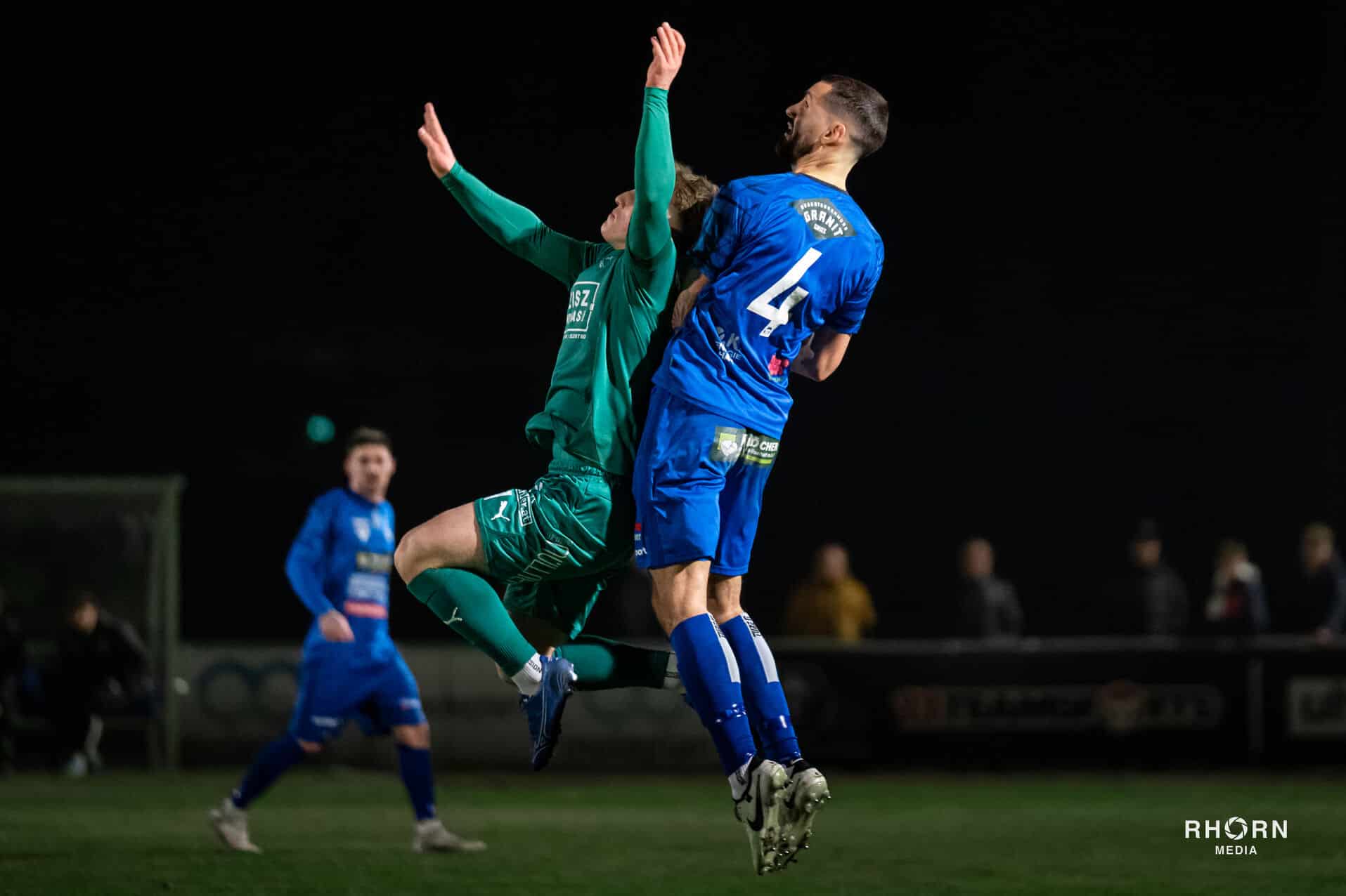Zwei Fußballspieler während eines Sprungduells im Flutlichtspiel, männlich, grüne und blaue Trikots.