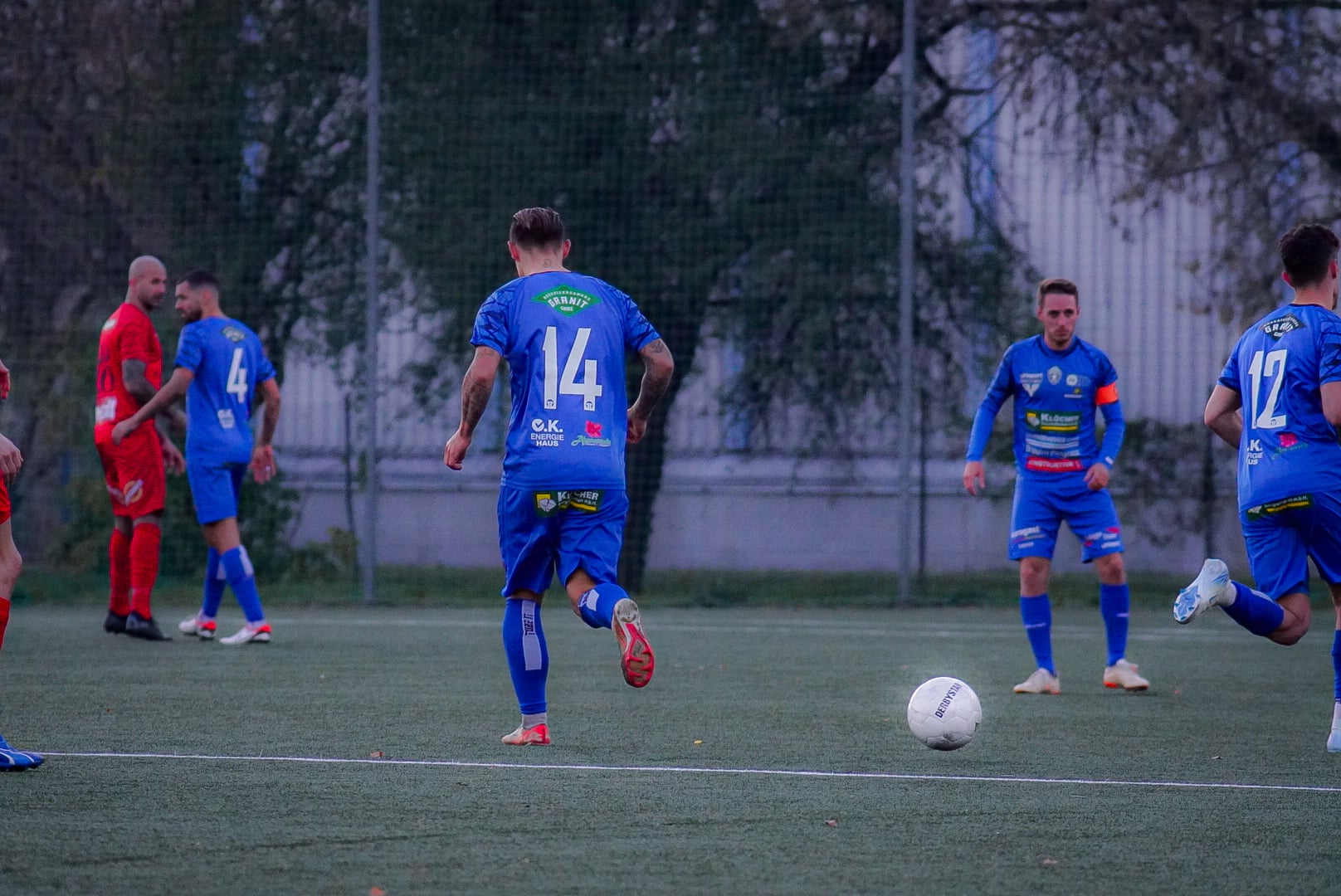 Fußballspiel auf dem Platz, Spieler in blauen Jerseys führen den Ball, Fokus auf Teamwork und Strategie im Spiel.