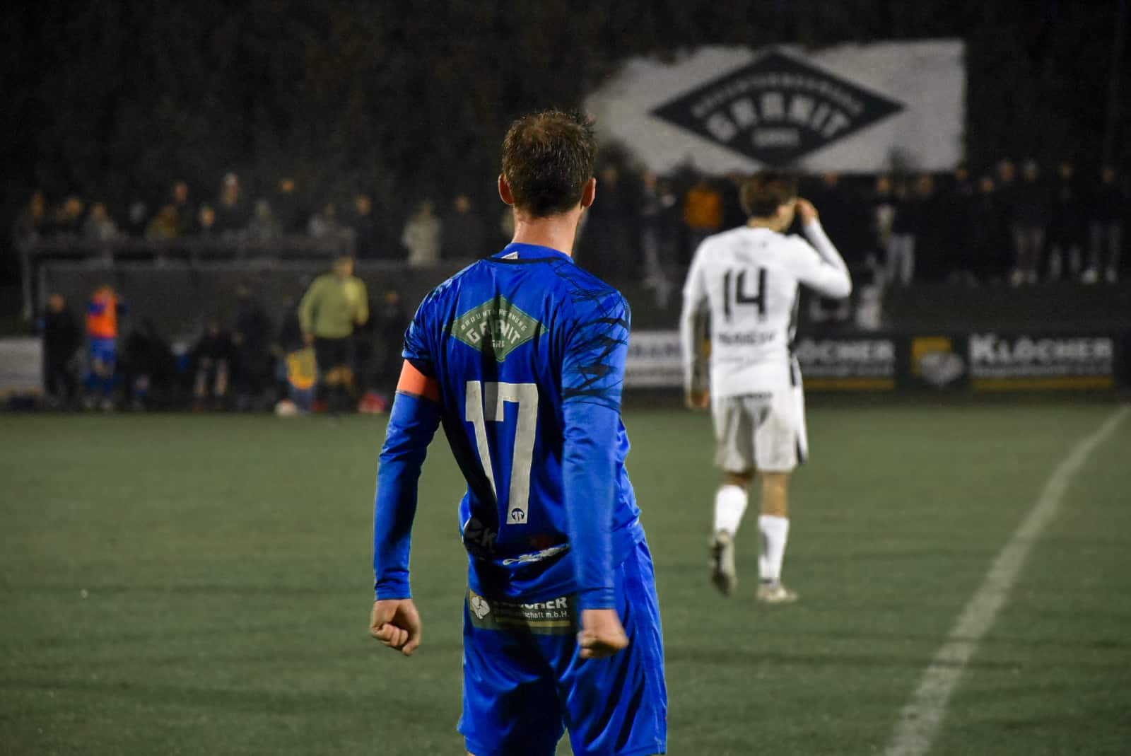 Fußballspieler im blauen Trikot mit Nummer 17 auf dem Spielfeld, Spieler in Weiß im Hintergrund, abendliches Spiel.