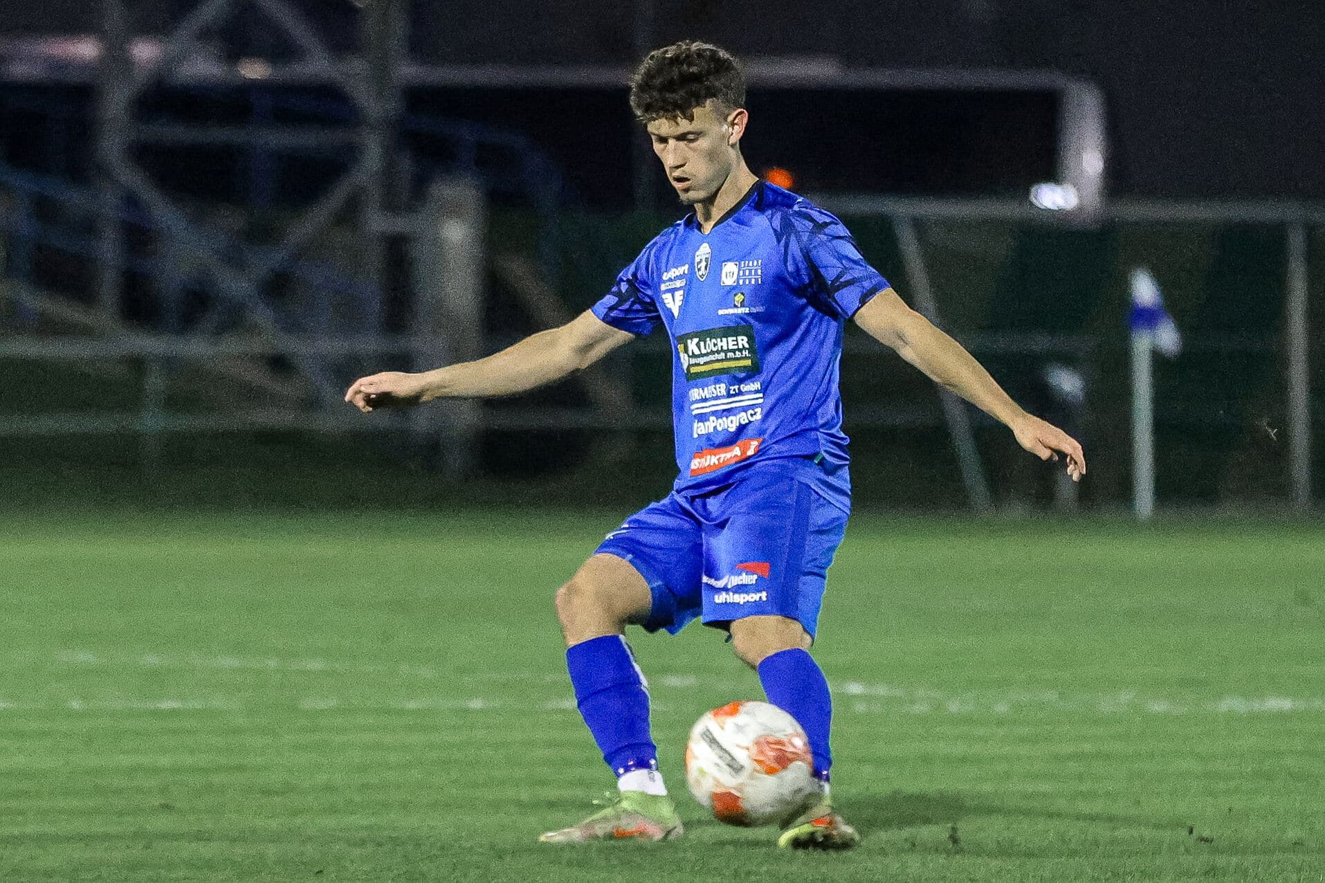 Fußballspieler in blauem Trikot kontrolliert den Ball auf dem Feld während eines Abendspiels.