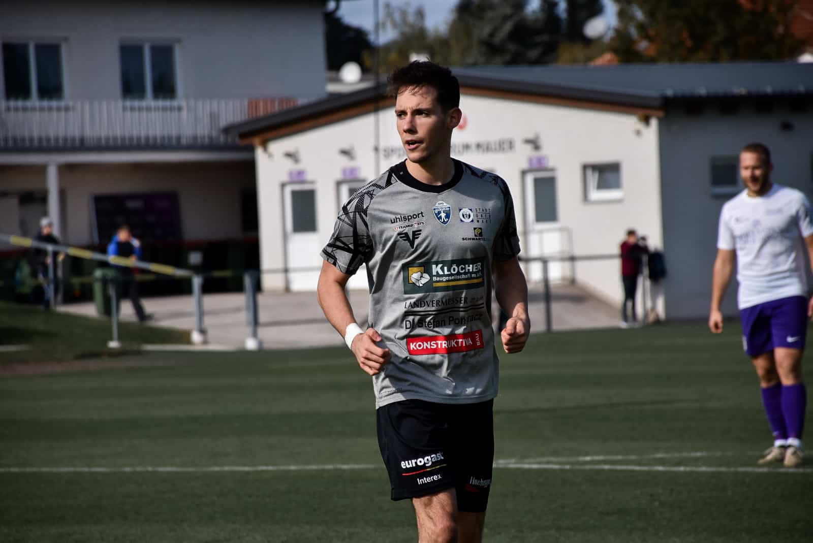 Ein Fußballspieler im grauen Trikot auf dem Spielfeld während eines Spiels, Zuschauer im Hintergrund.