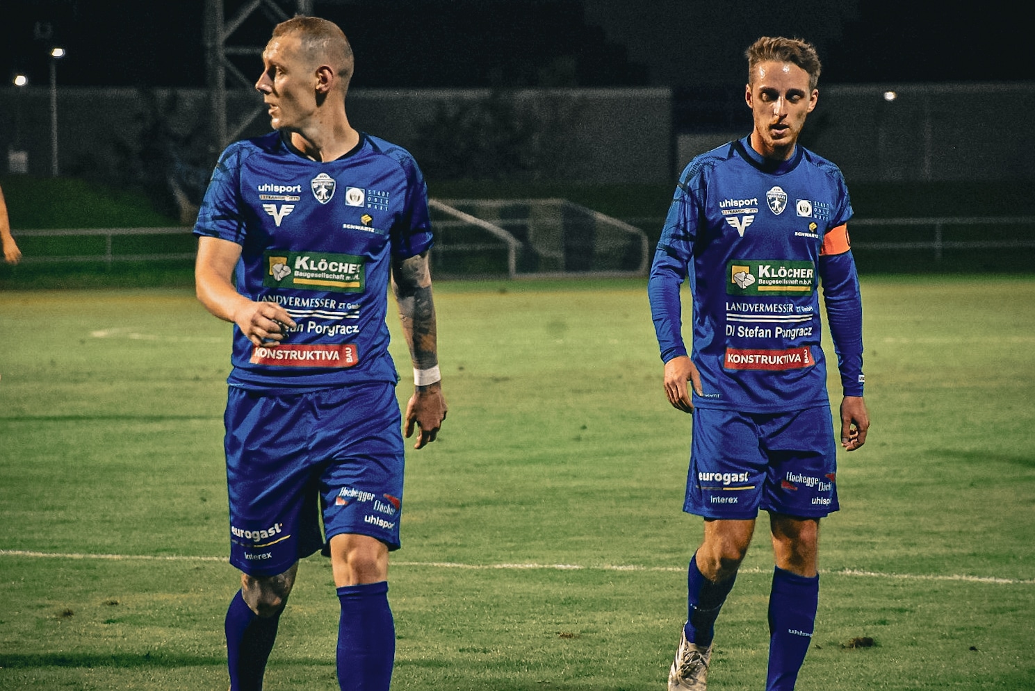 Zwei Fußballspieler in blauen Trikots auf dem Spielfeld bei Nacht, bereit für das Spiel.