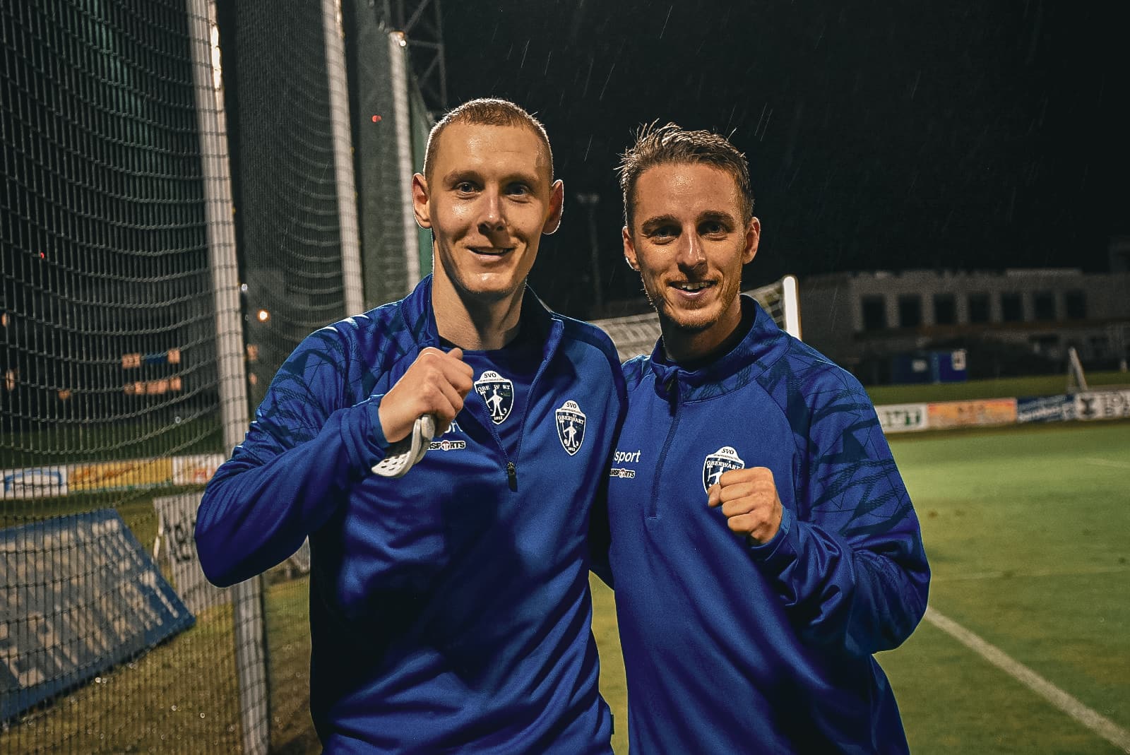 Zwei Fußballspieler im blauen Trikot feiern auf dem Spielfeld bei Nacht, lächelnd und mit erhobener Faust.