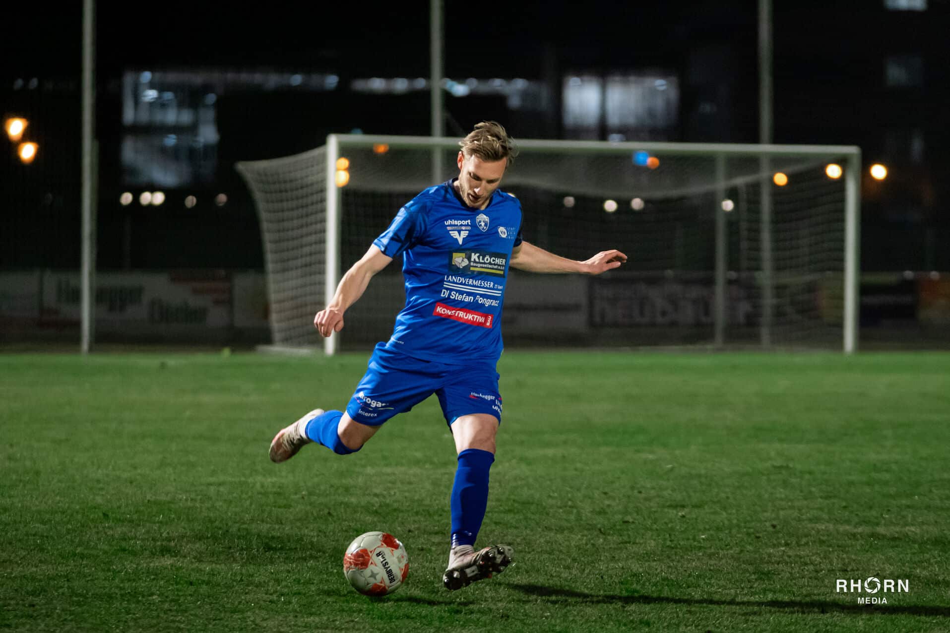 Ein Fußballspieler im blauen Trikot schießt im nächtlichen Stadion Richtung Tor.