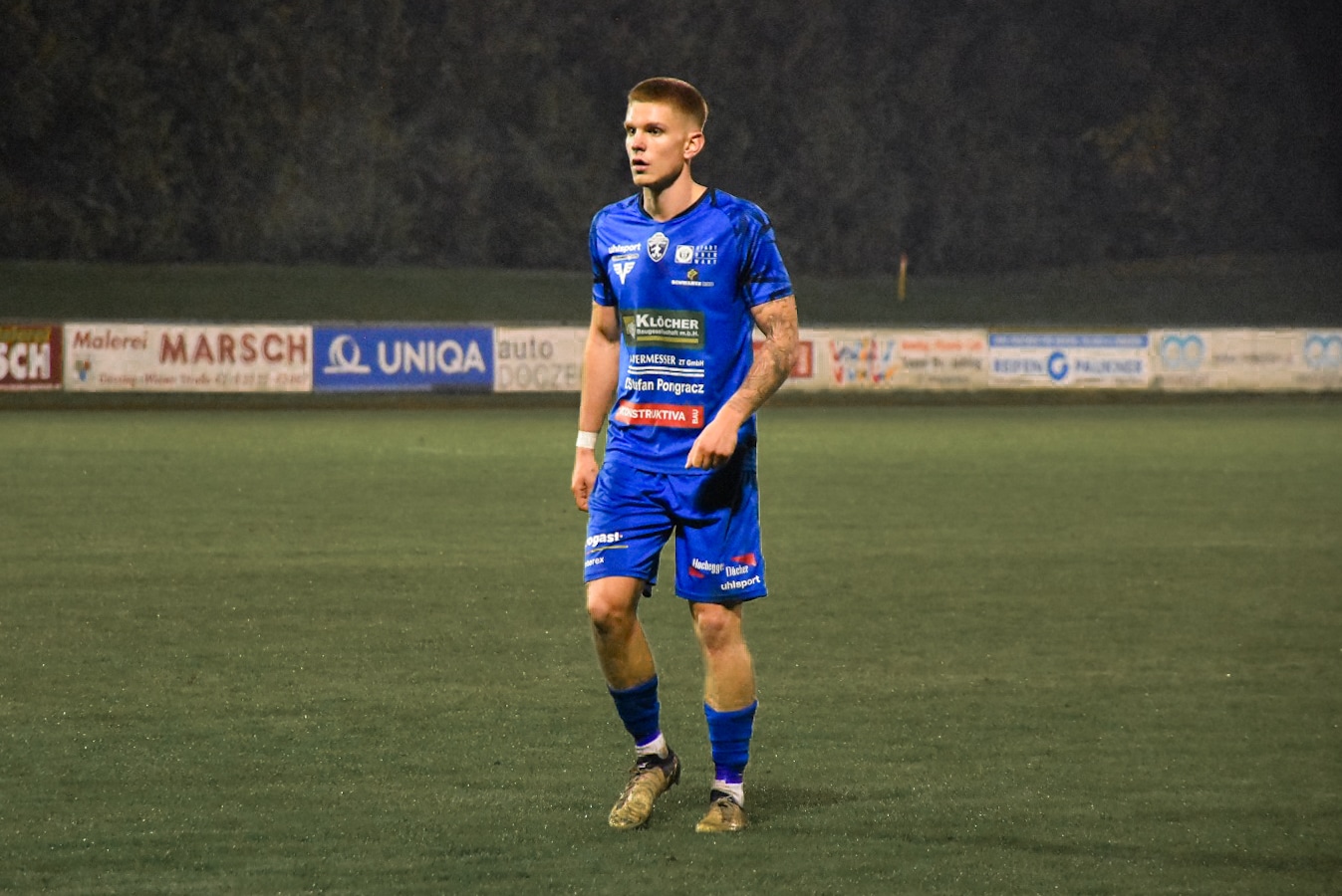 Fußballspieler im blauen Trikot auf dem Spielfeld bei Nachtspiel, umgeben von Werbebannern.