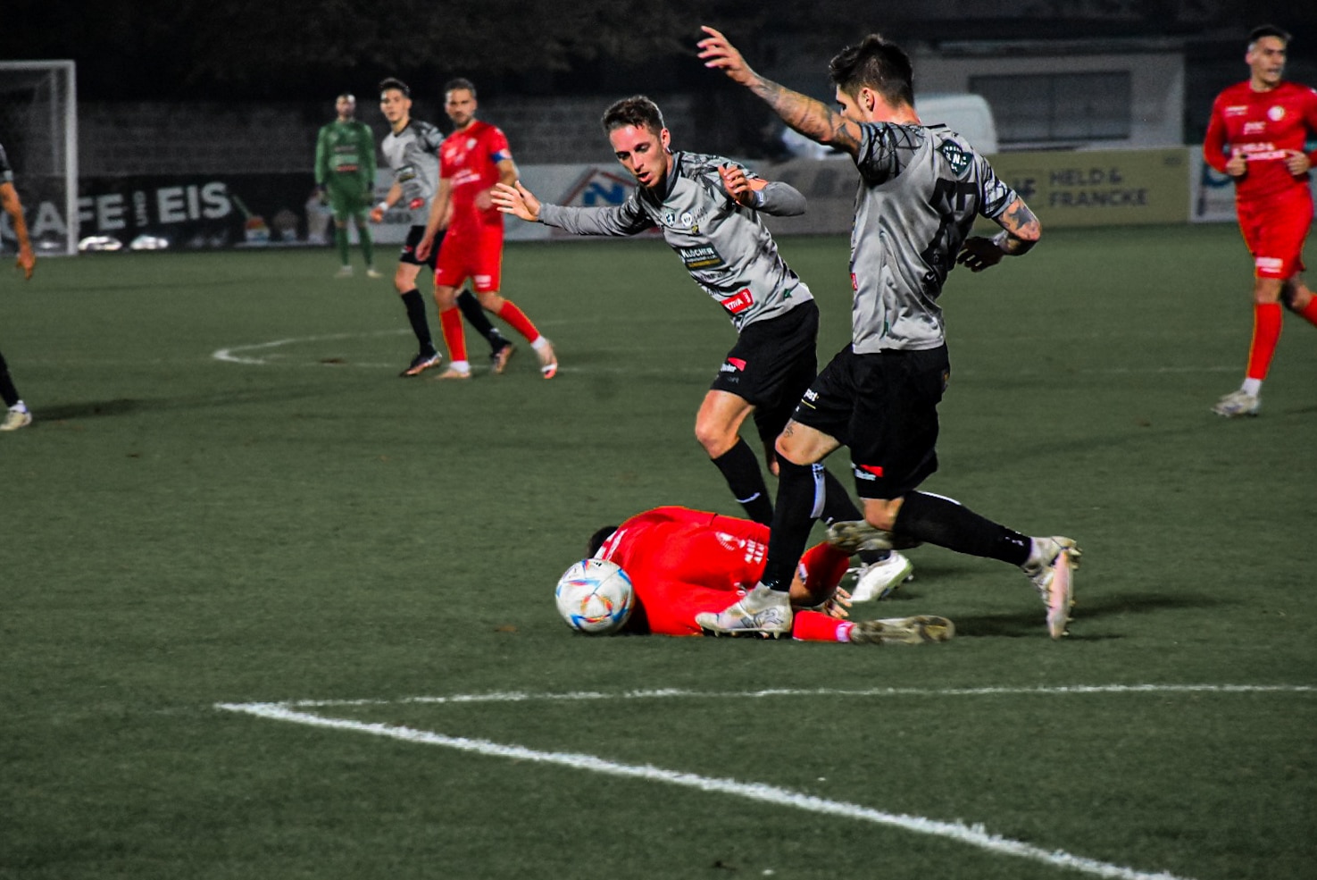 Fußballspielaktion auf dem Feld, Spieler in roten und grauen Trikots beim Kampf um den Ball, gestikulierend und fallend.