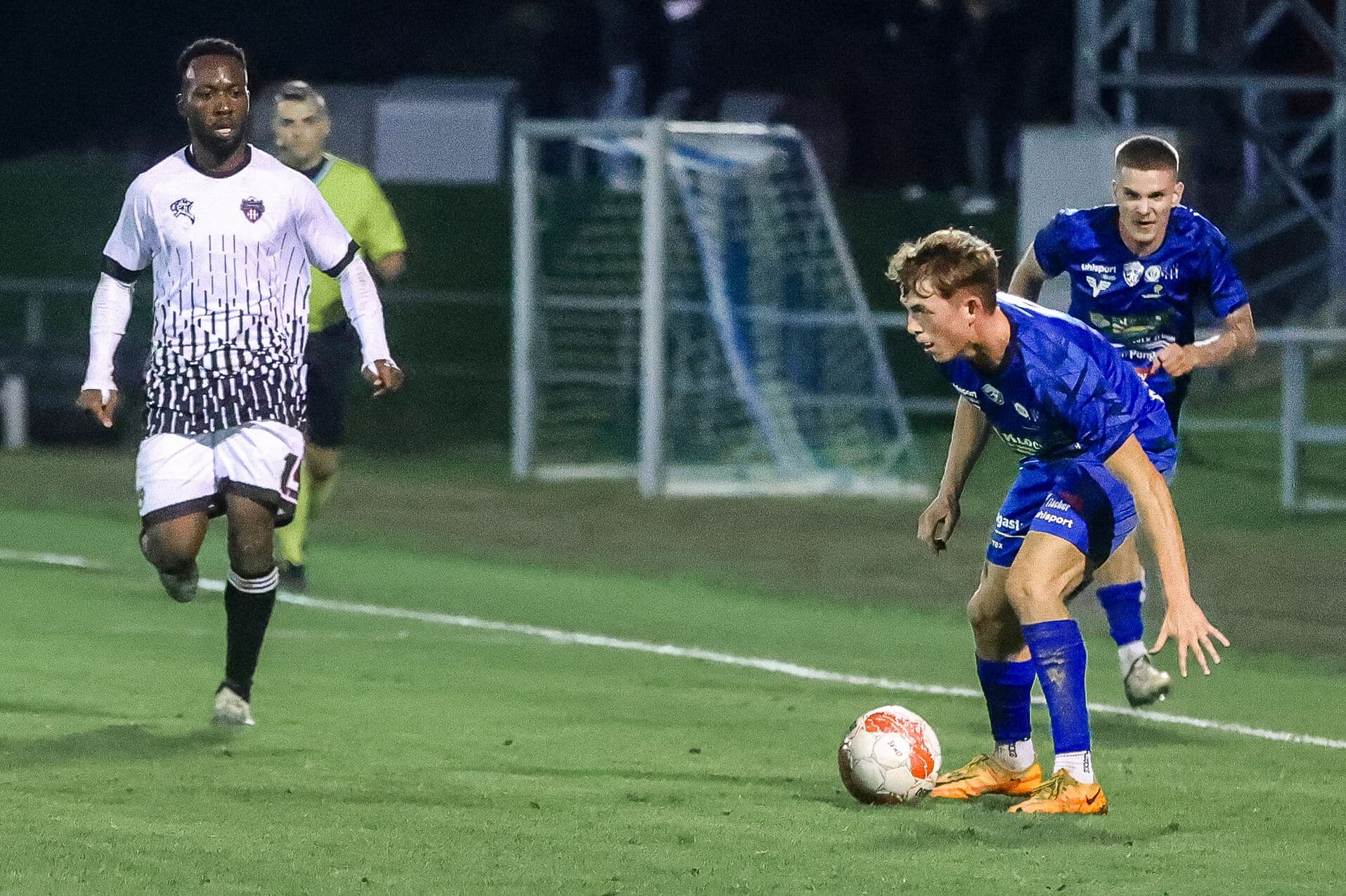 Fußballspieler in Aktion auf dem Feld bei Nachtspiel, blaues und weißes Team, nah am Tor.
