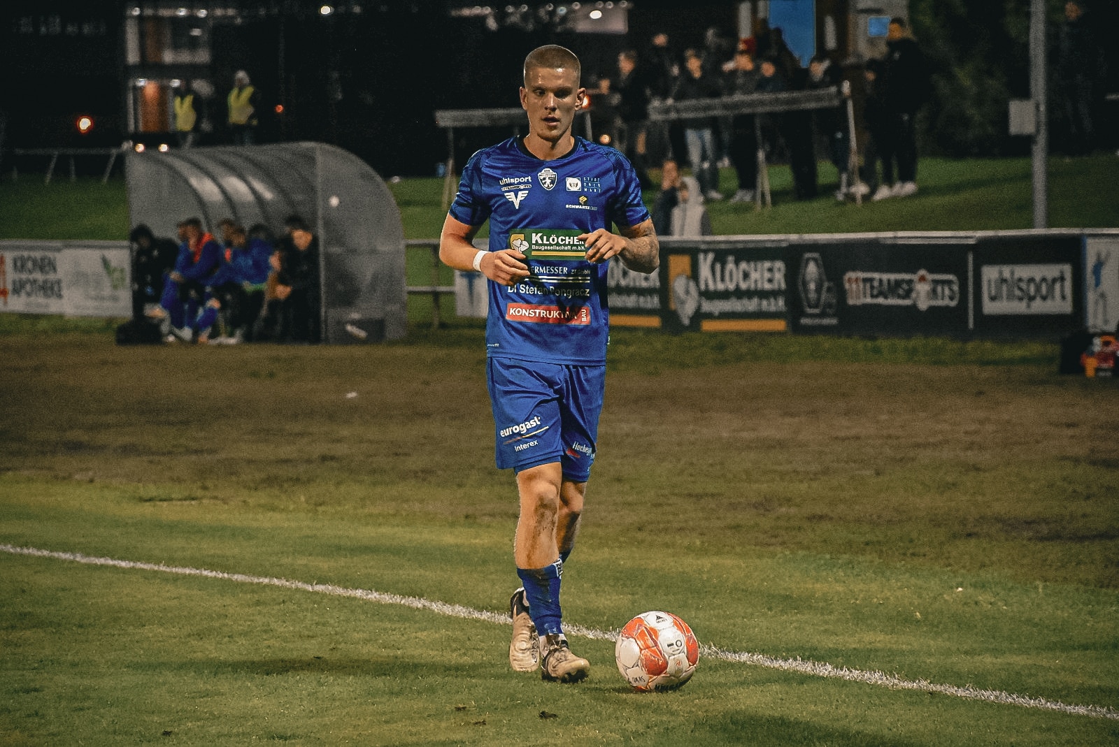 Fußballspieler in blauem Trikot bei Nacht, bereitet sich auf einen Einwurf vor, Spielfeld und Zuschauer im Hintergrund.
