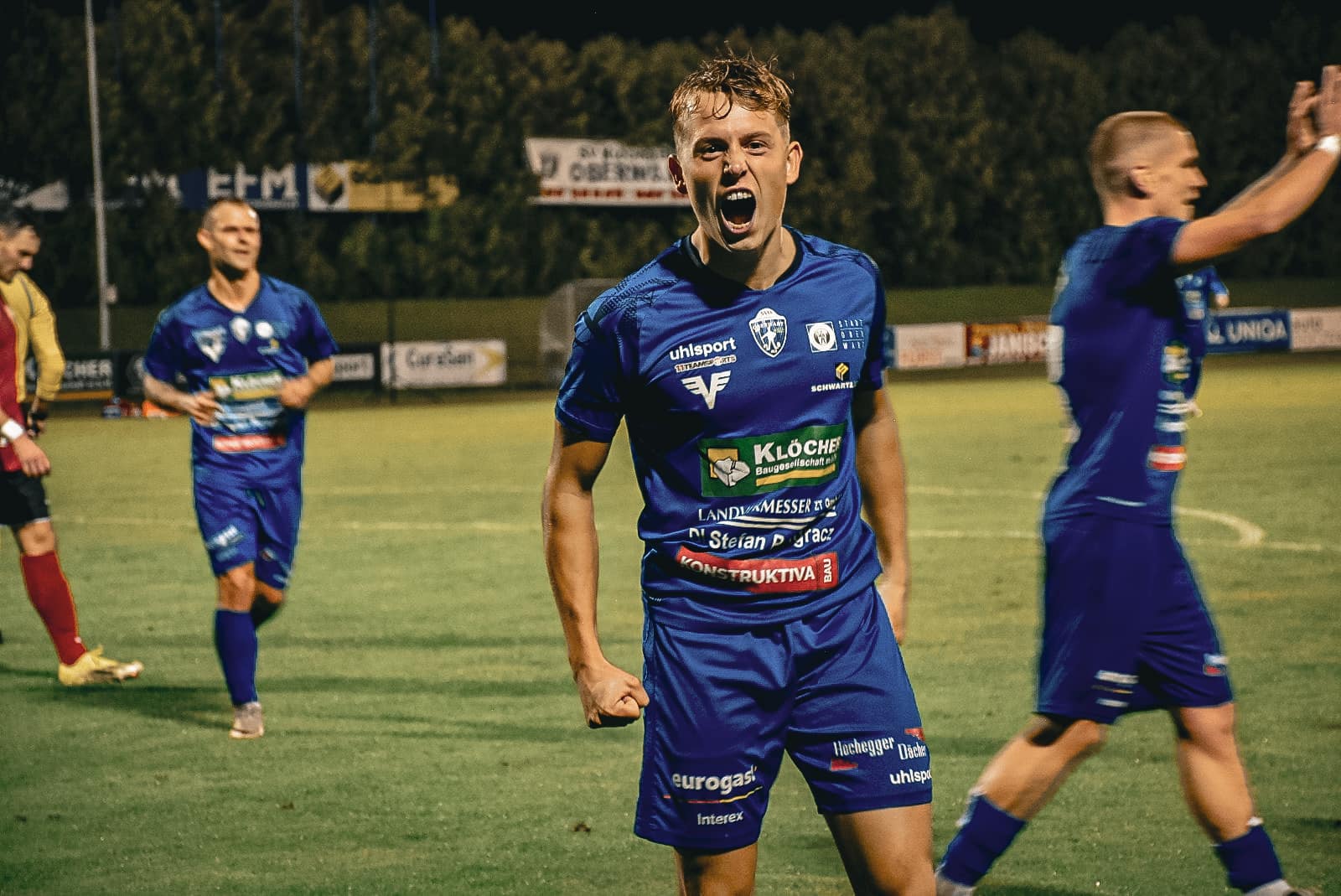 Ein jubelnder Fußballspieler im blauen Trikot feiert auf dem Spielfeld unter Flutlicht.