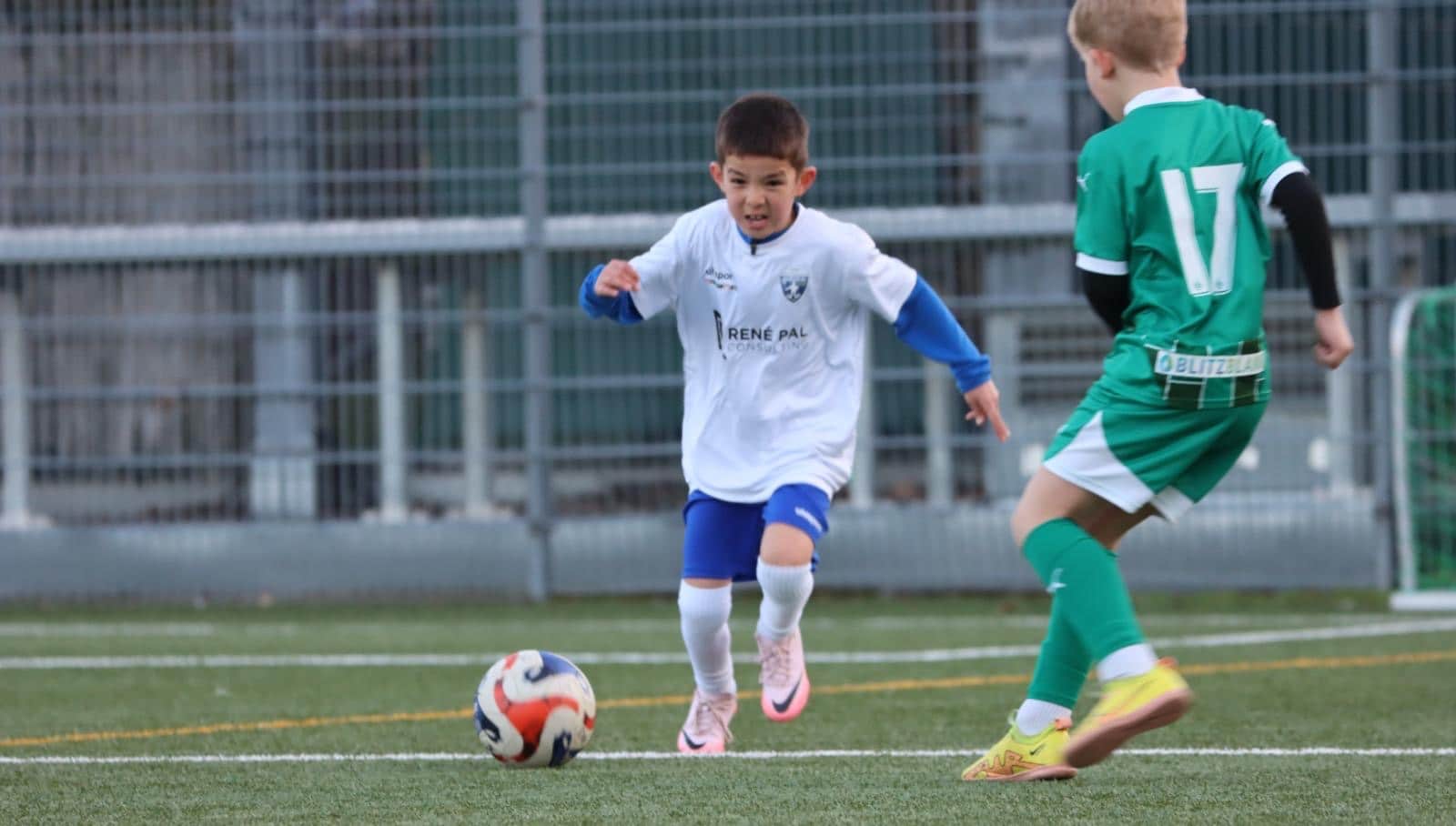 Zwei junge Fußballer im Zweikampf auf dem Spielfeld, Spieler in Weiß dribbelt gegen Spieler in Grün.