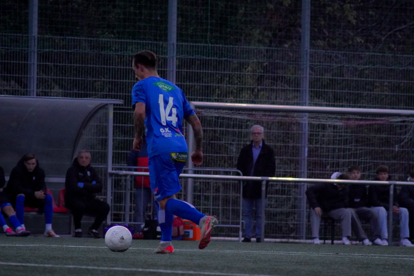 Ein Fußballspieler in blauem Trikot, Nummer 14, auf einem Kunstrasenfeld. Zuschauer und Ersatzbank im Hintergrund.
