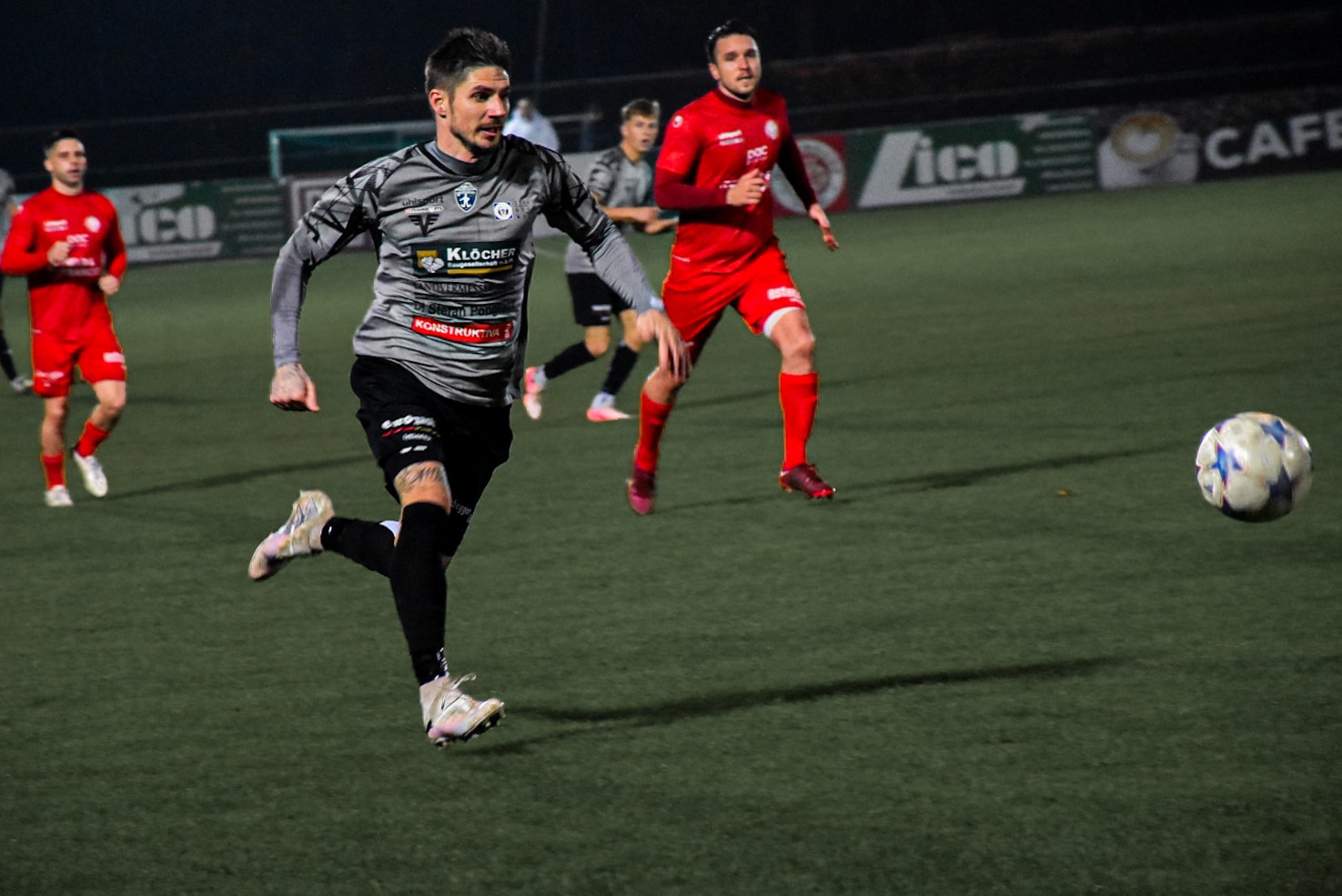 Fußballspieler im grauen Trikot rennt auf dem Spielfeld, verfolgt den Ball im spannenden Match gegen das rote Team.