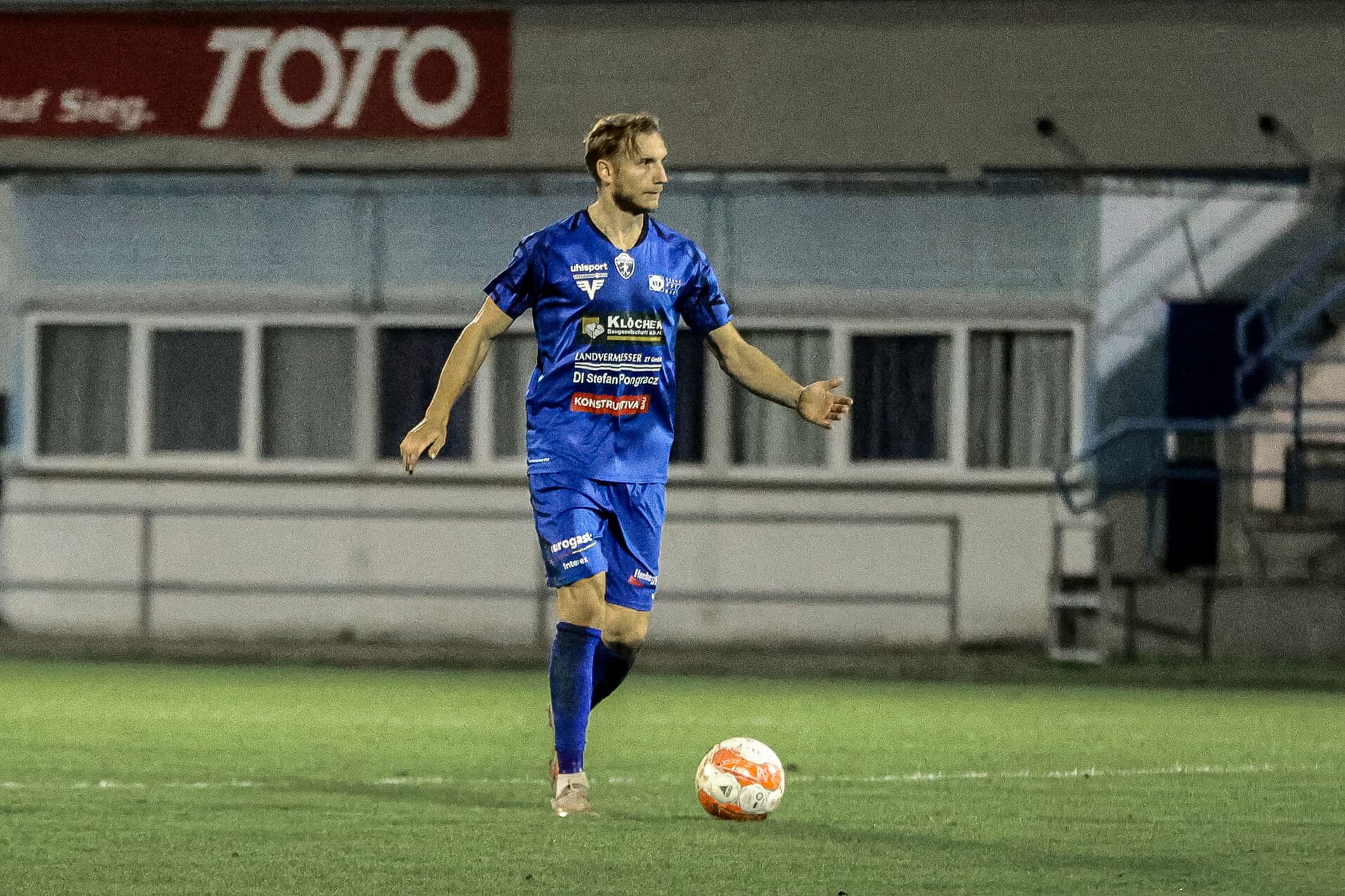 Fußballspieler in blauem Trikot kontrolliert den Ball auf dem Spielfeld während eines Nachspiels.