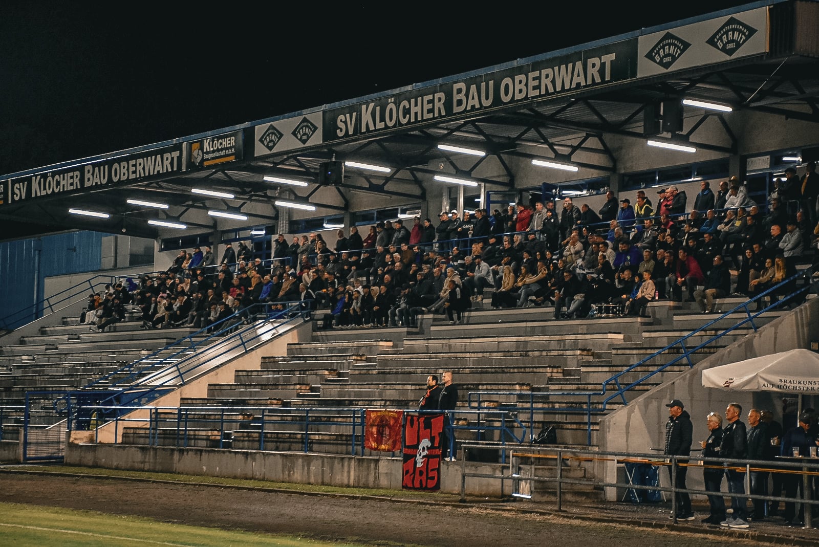 Zuschauer auf Tribüne beim SV Klöcher Bau Oberwart Fußballspiel am Abend, Stadionansicht.