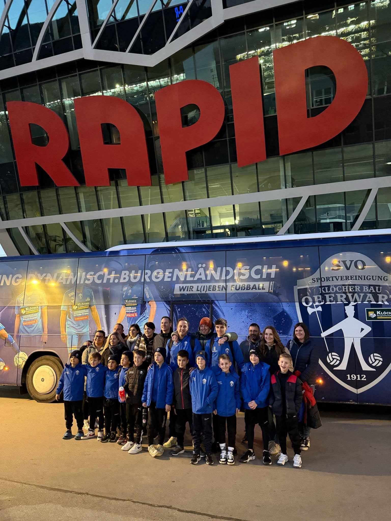 Gruppe vor Rapid-Stadion mit Team-Bus, lächelnd in Sportkleidung. Freude beim Fußballabend in Wien.