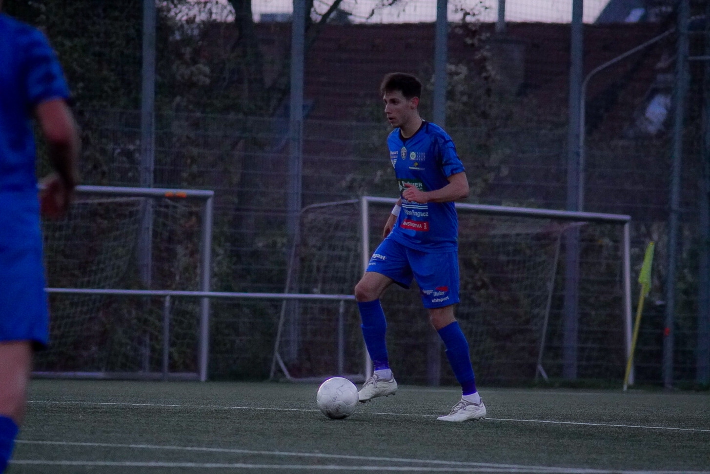 Fußballspieler in blauem Trikot dribbelt den Ball auf einem Spielfeld vor dem Tor bei Abenddämmerung.