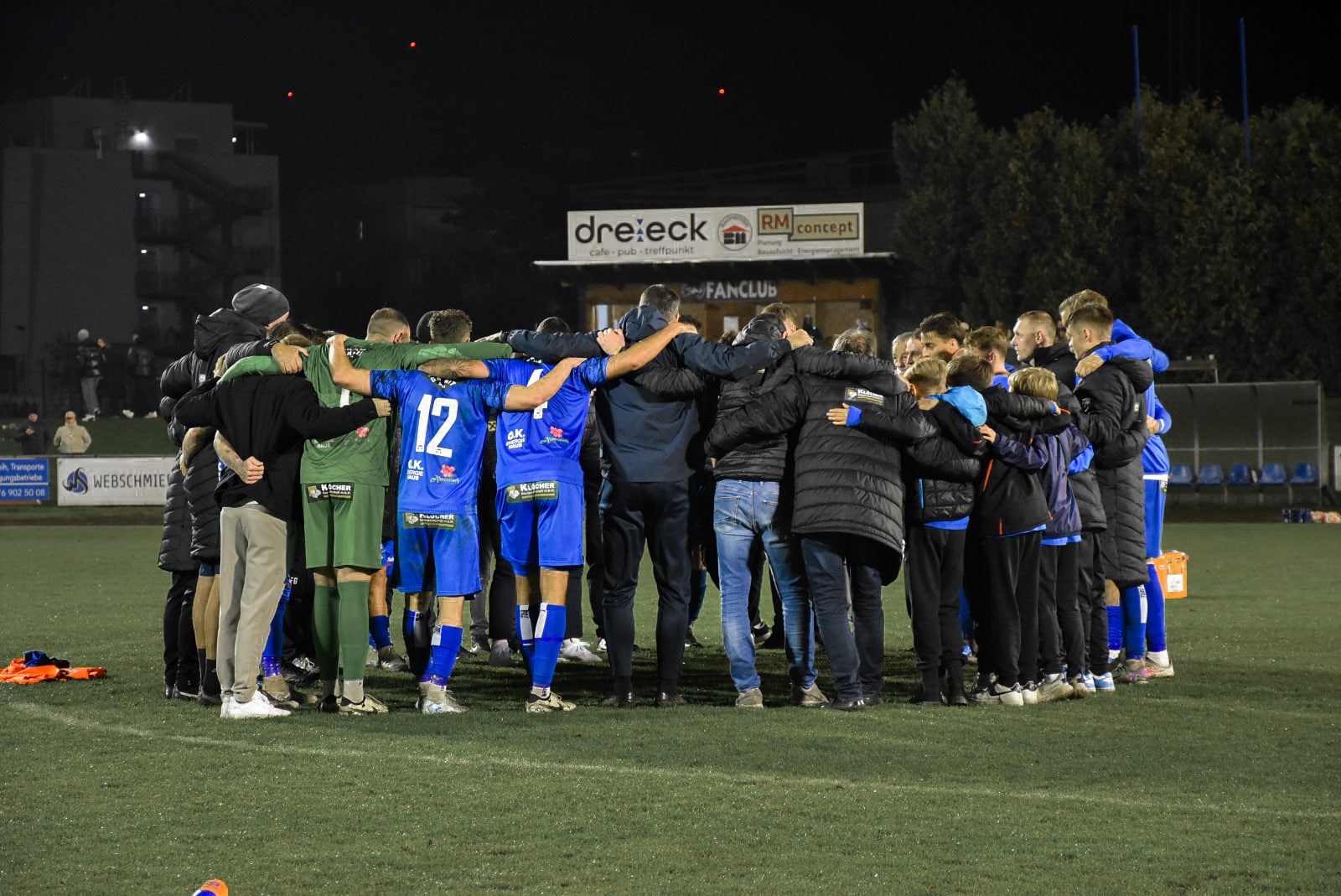 Fußballteamkreis bei Nacht auf dem Spielfeld, Spieler in blauen Trikots, gemeinsamer Teamgeist.