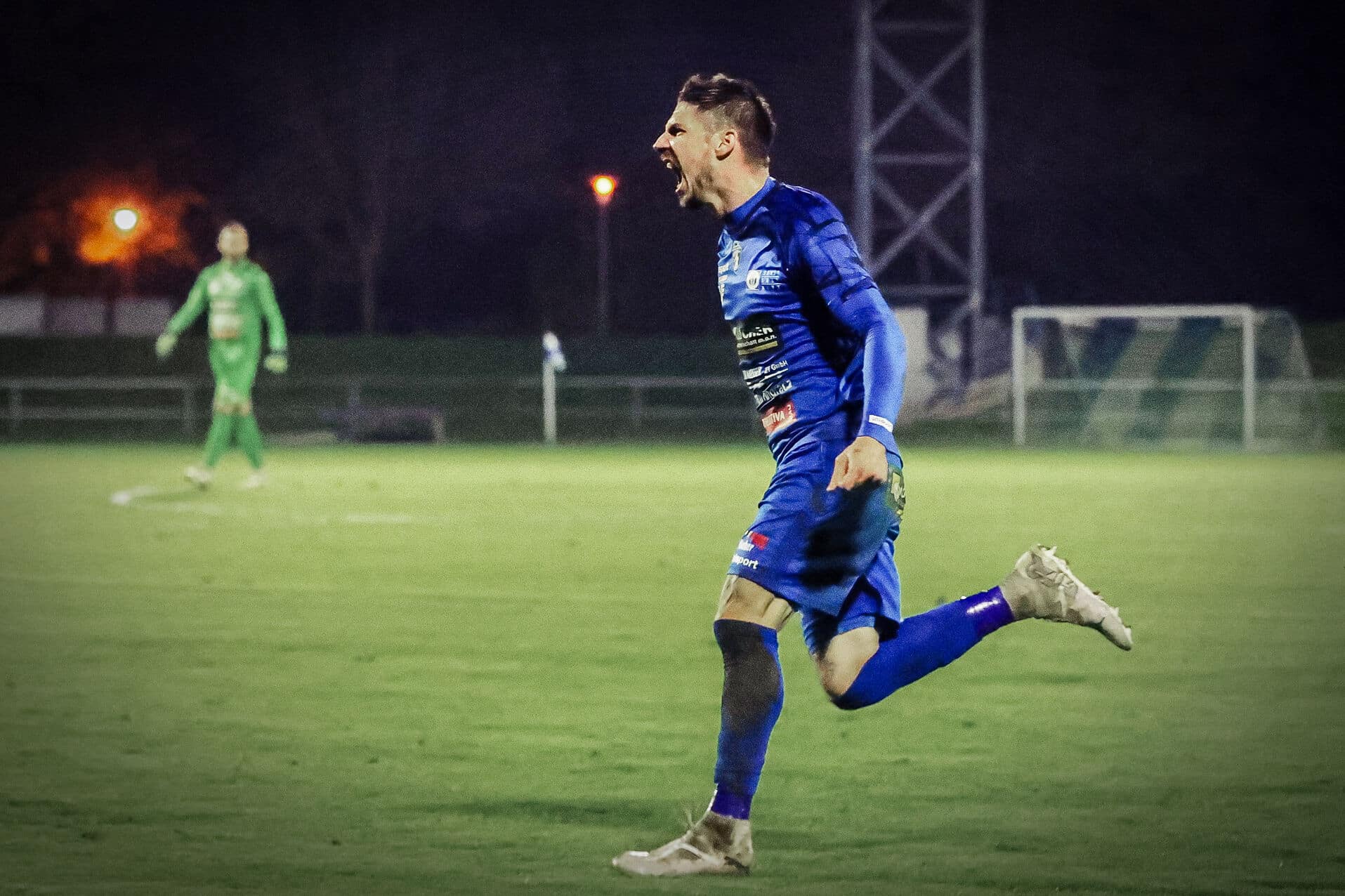 Fußballspieler jubelt auf dem Spielfeld bei Nachtspiel, im blauen Trikot, ausdrucksstarker Torjubel.