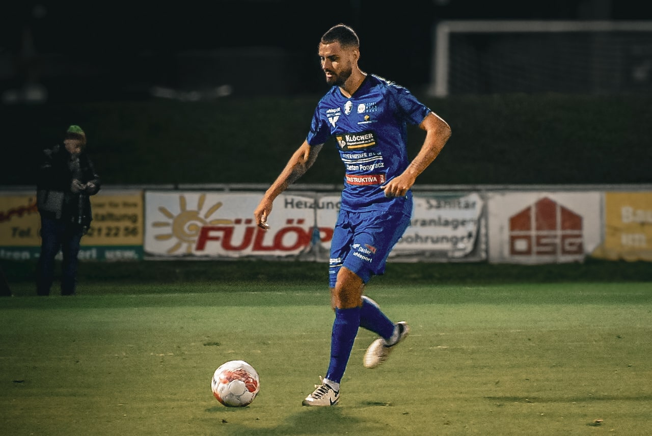 Fußballspieler im blauen Trikot dribbelt den Ball bei einem Abendspiel auf dem Spielfeld.