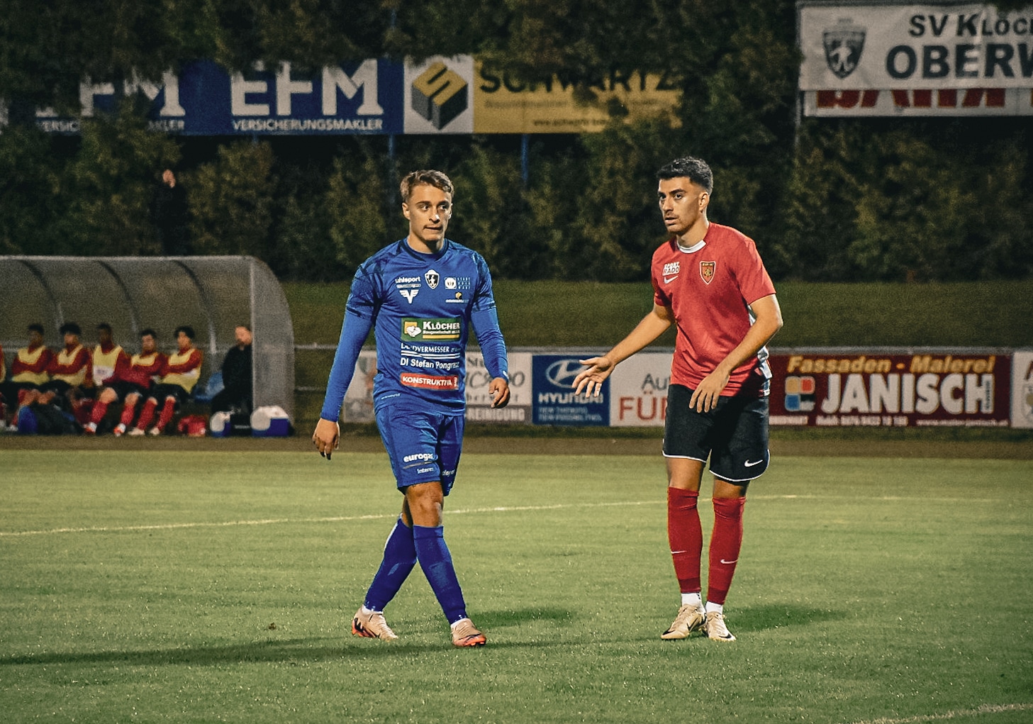 Zwei Fußballspieler im blauen und roten Trikot während eines Nachtspiels auf dem Spielfeld.