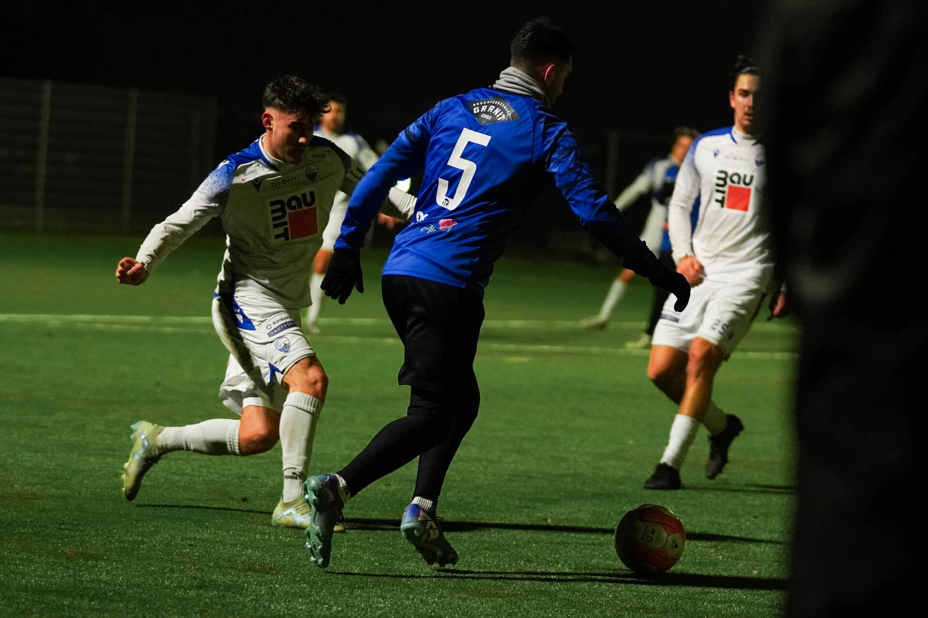 Fußballspiel bei Nacht: Spieler in blauen und weißen Trikots im intensiven Zweikampf um den Ball auf dem Spielfeld.
