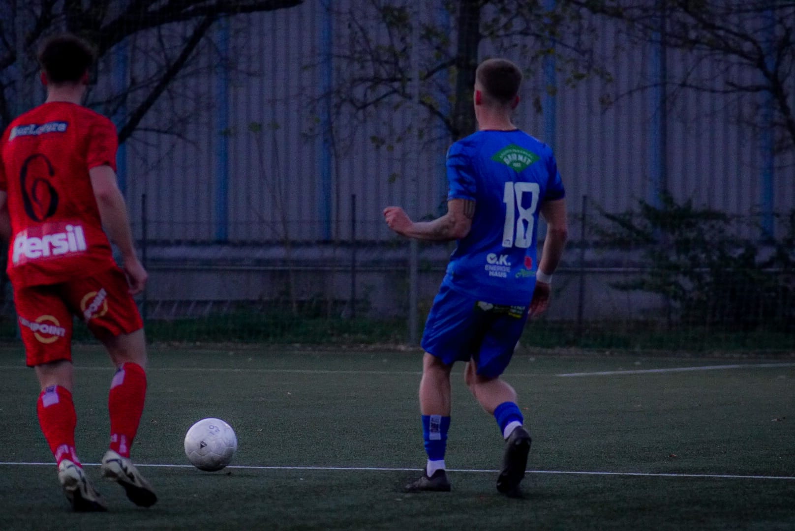 Zwei Fußballspieler in roten und blauen Trikots beim Spiel auf einem grünen Platz mit einem weißen Ball.