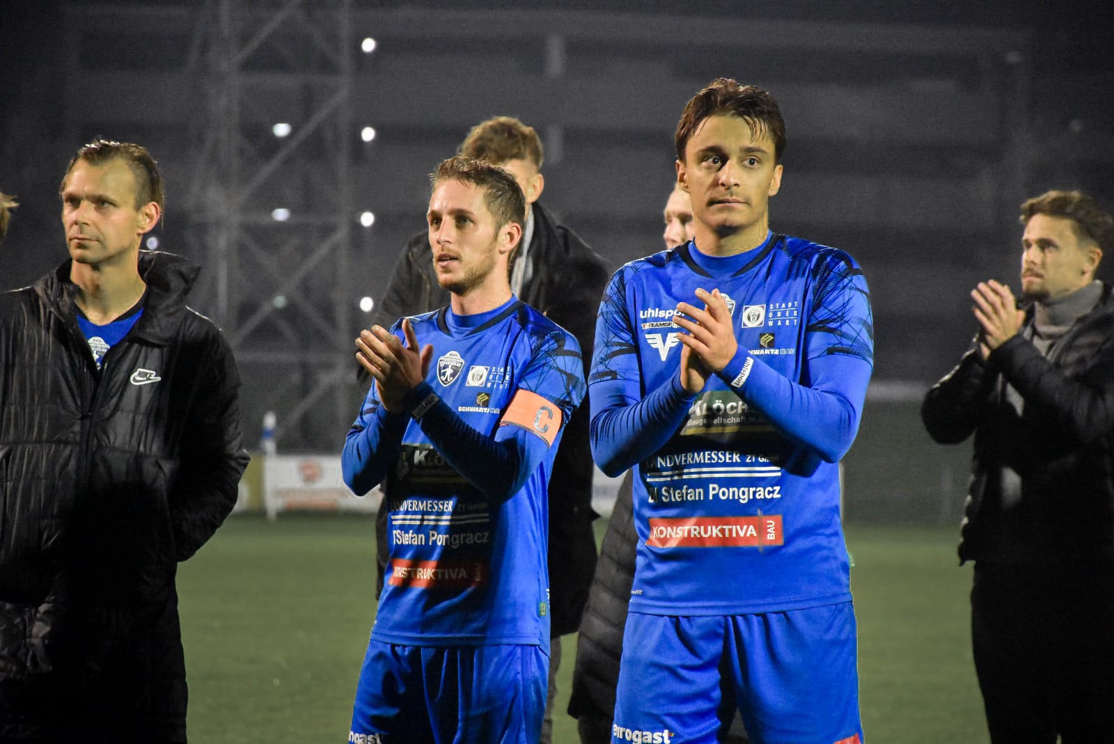 Fußballspieler klatschen nach Spiel auf dem Feld, tragen blaue Trikots mit Sponsorenlogos, Abendspielbeleuchtung.