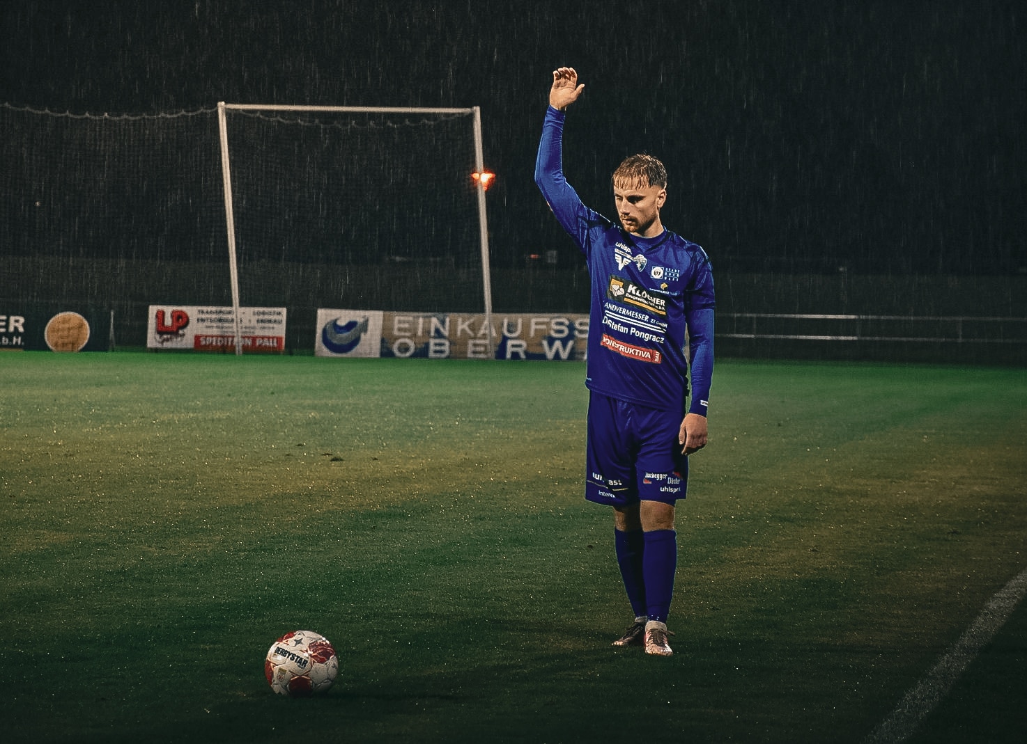 Fußballspieler im blauen Trikot bei Regen auf dem Spielfeld beim Freistoßabendspiel, hebt den Arm.