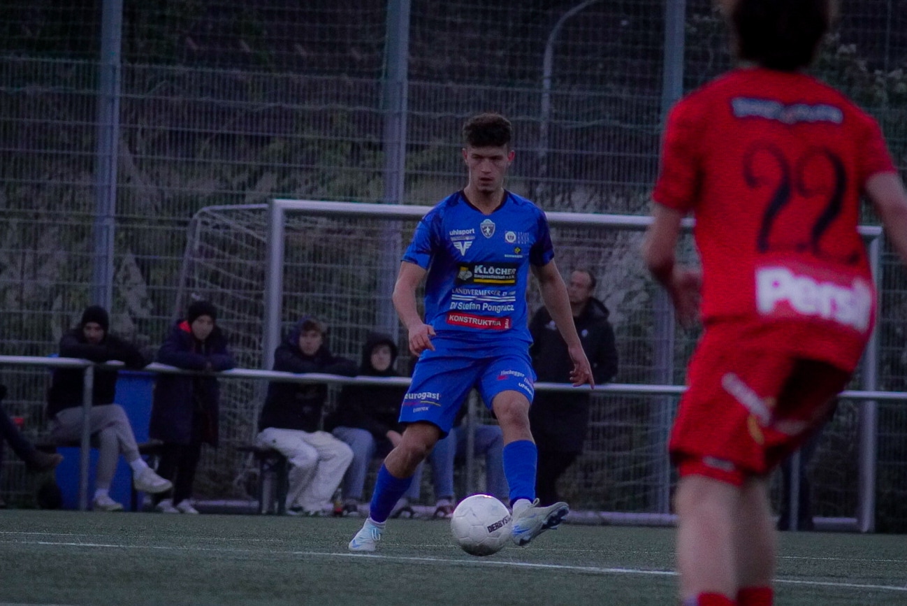 Fußballspieler in blauem Trikot kontrolliert den Ball auf dem Spielfeld, während Zuschauer am Spielfeldrand zuschauen.