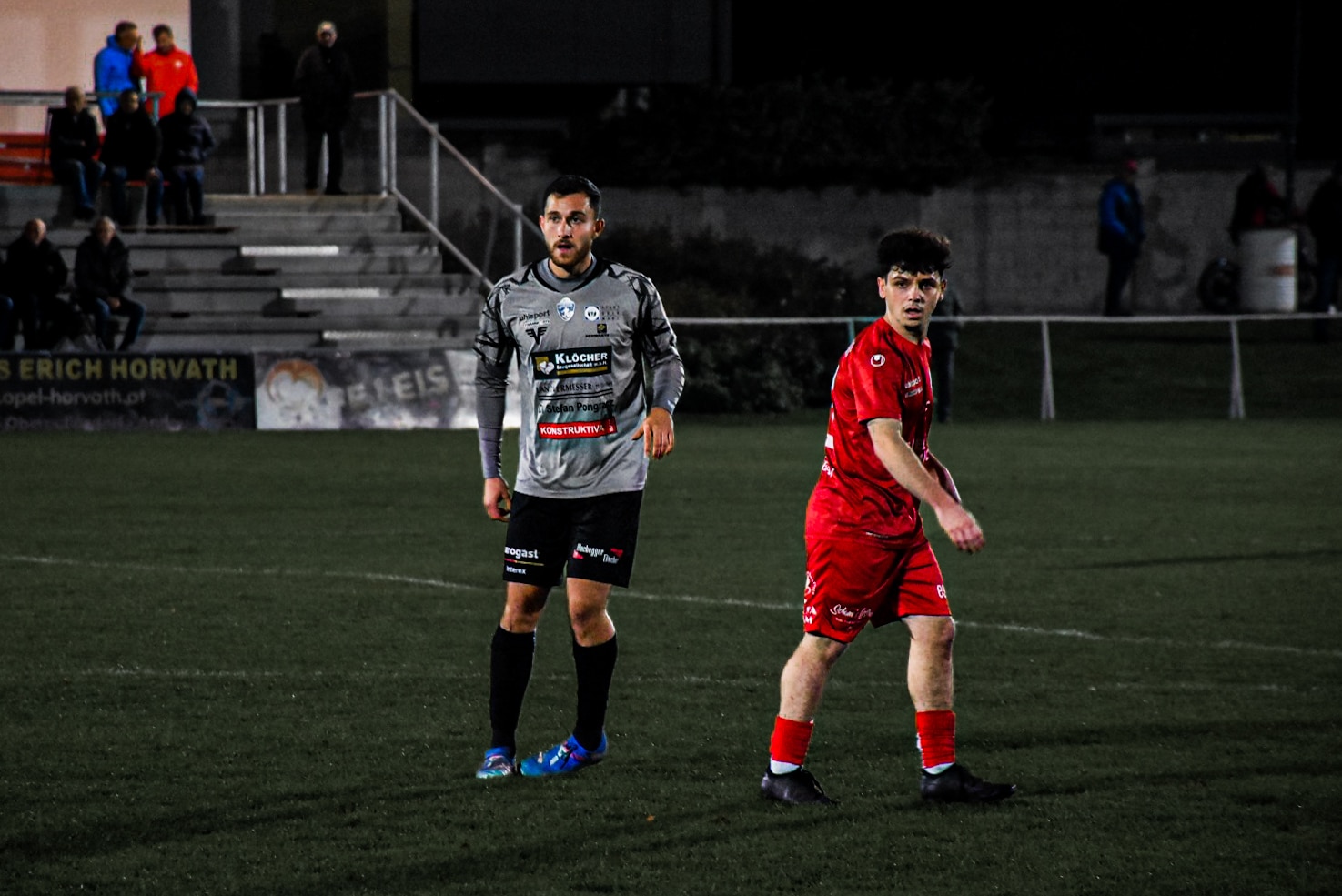 Fußballspieler im Spiel bei Nacht auf dem Feld, mit Zuschauern im Hintergrund auf der Tribüne.