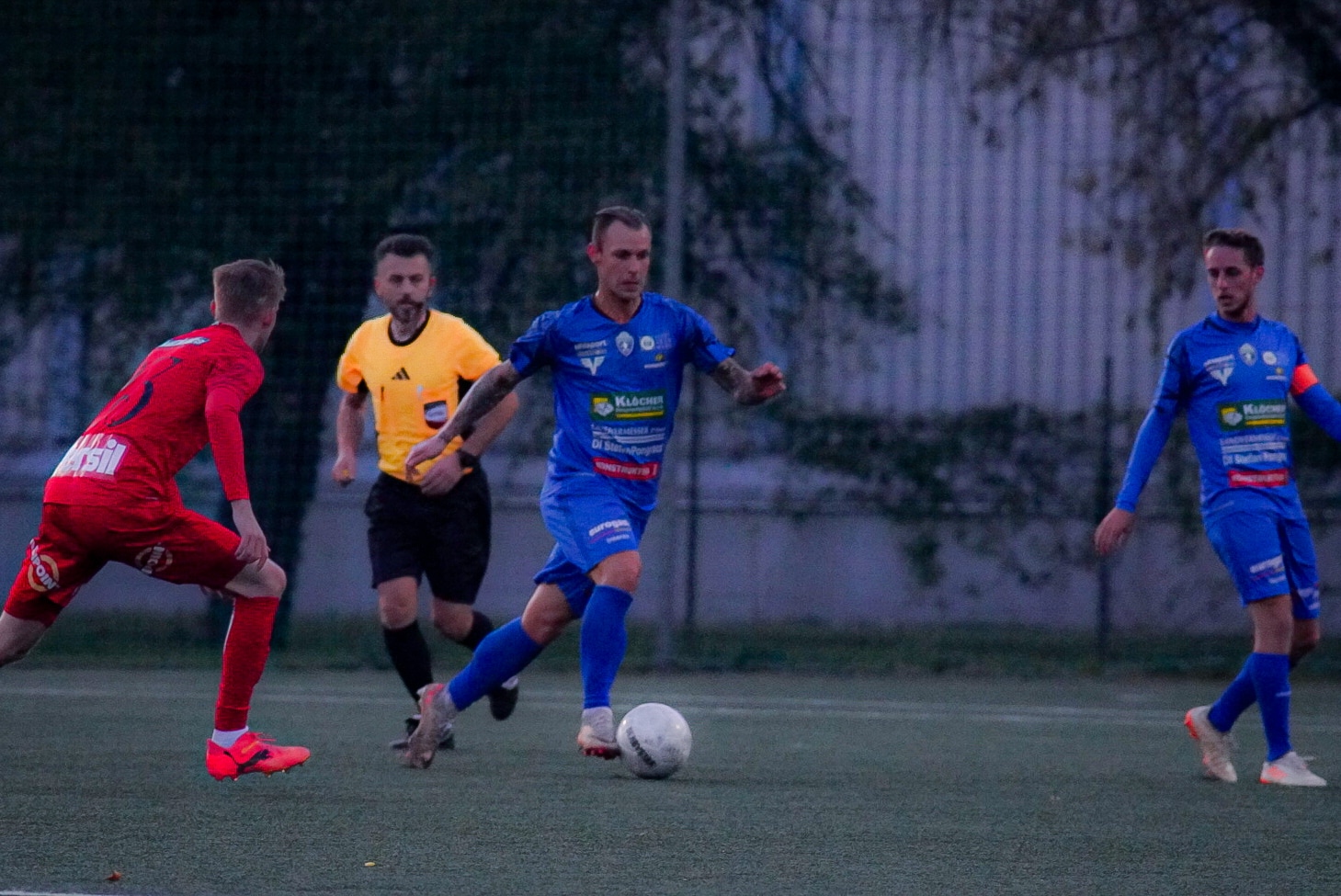 Fußballspiel: Spieler in Blau dribbelt Ball, verfolgt von Gegenspielern in Rot, Schiedsrichter nah am Geschehen.