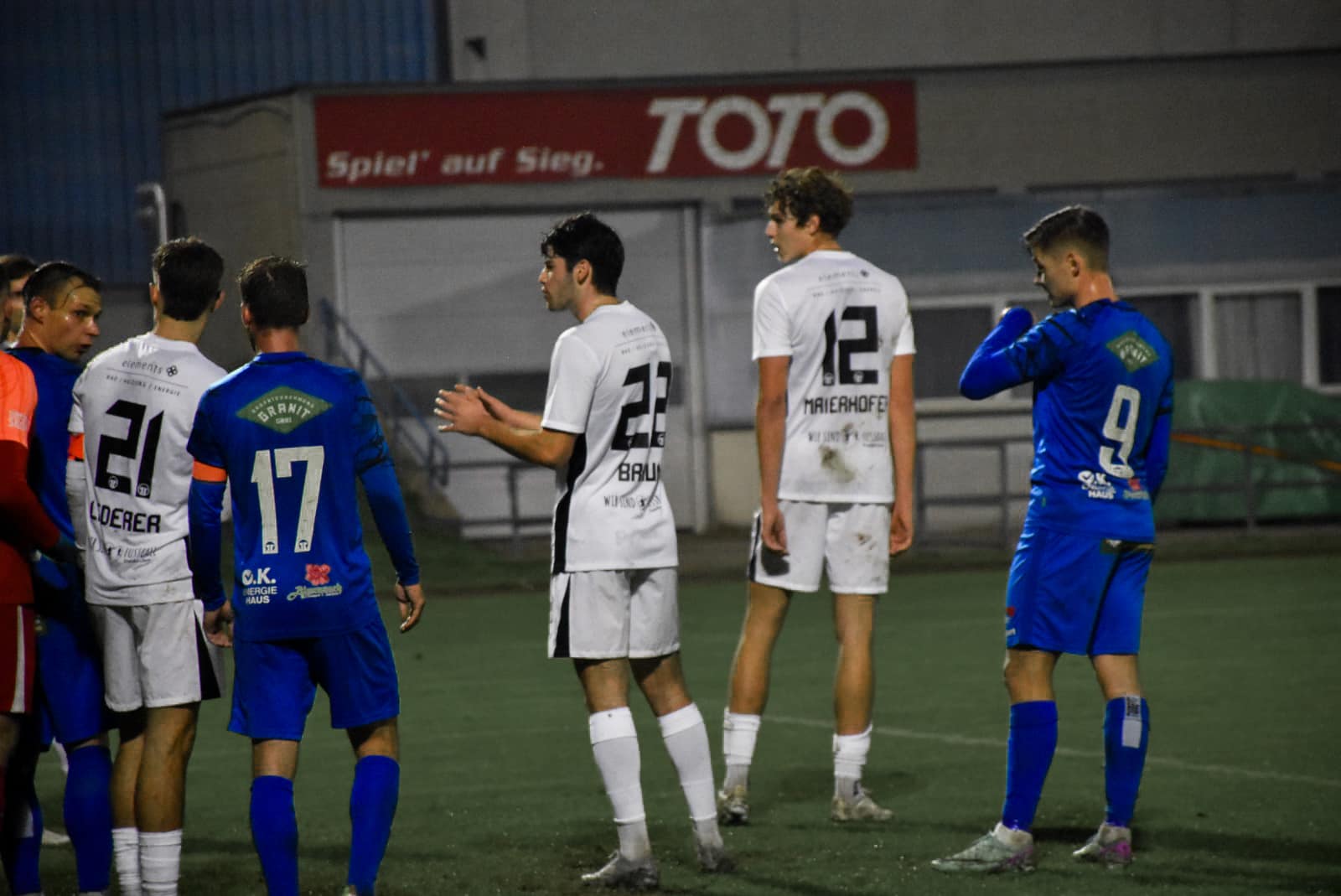 Fußballspiel bei Nacht, Spieler in blau-weißen Trikots diskutieren auf dem Spielfeld unter „TOTO“-Banner.