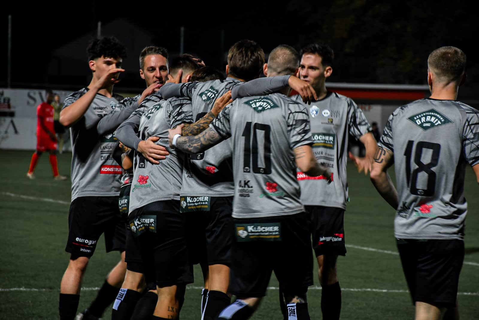 Fußballteam feiert Tor auf dem Spielfeld, Spieler in grauen Trikots, Nachtspiel, sportliche Begeisterung und Teamgeist.