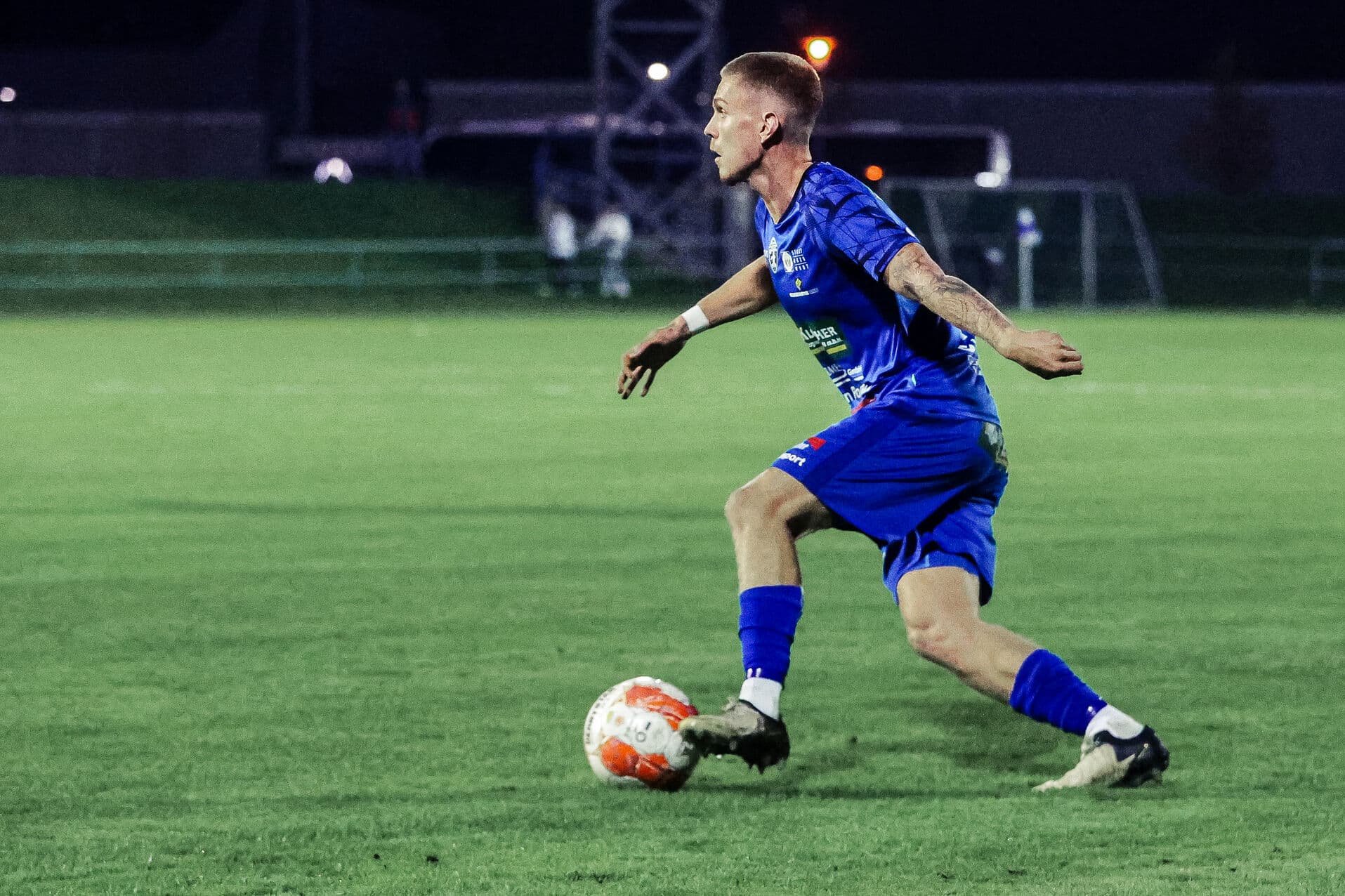 Fußballspieler in blauer Uniform auf dem Spielfeld beim Dribbling mit einem orange-weißen Ball bei Nacht.