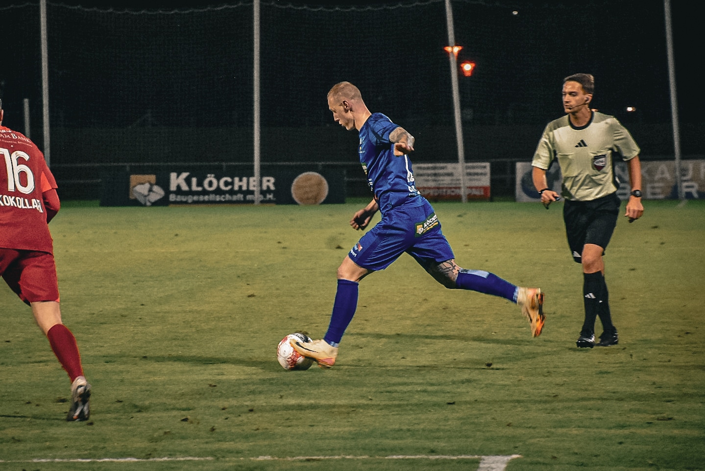 Fußballspieler in blauem Trikot dribbelt den Ball in einem nächtlichen Spiel auf dem Spielfeld.