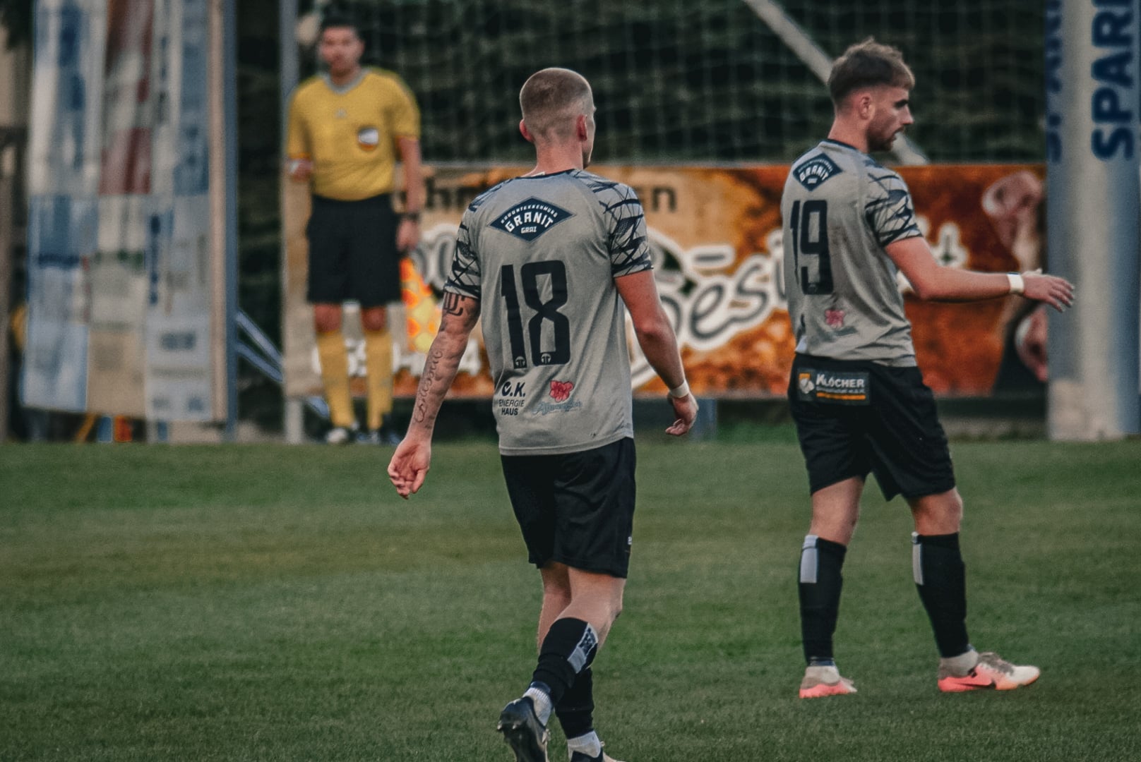 Zwei Fußballspieler in grauen Trikots auf dem Spielfeld, Schiedsrichter im Hintergrund, bereit zum Spiel.