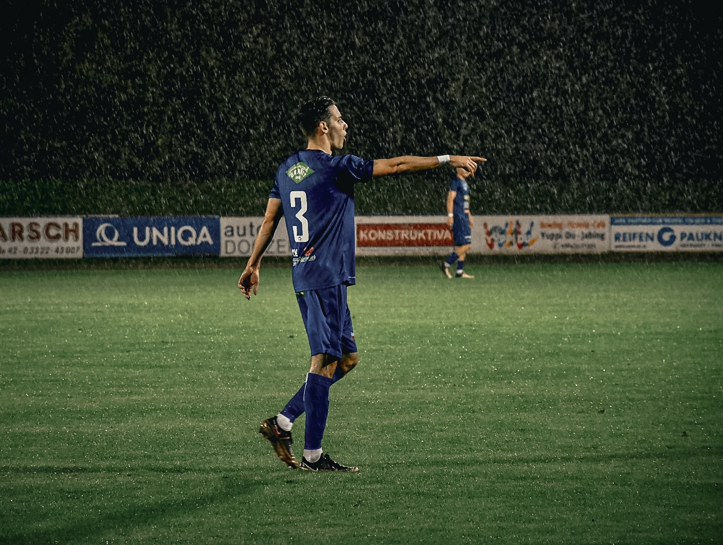 Fußballspieler in blauem Trikot spielt bei Regen auf dem Feld und zeigt mit dem Finger in eine Richtung.