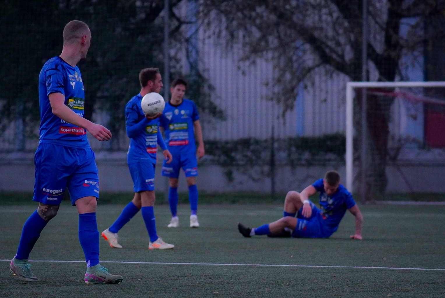 Fußballspieler in blauen Trikots auf dem Spielfeld, einer sitzt am Boden, andere stehen mit Ball in der Hand.