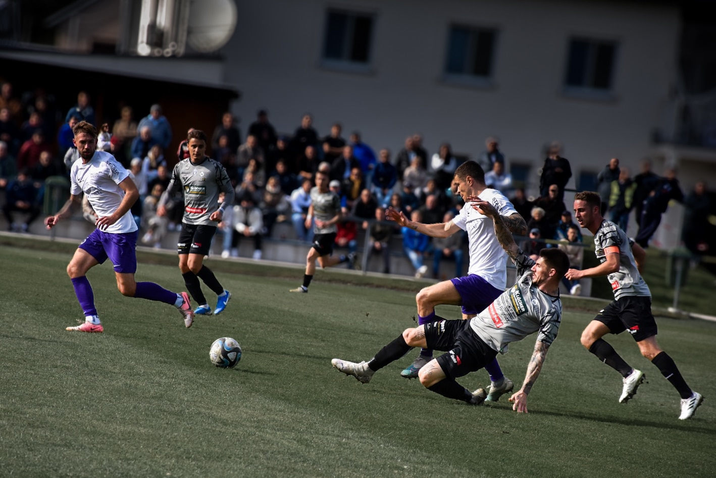 Fußballspiel in Aktion: Spieler im Zweikampf um den Ball auf dem Feld, umgeben von Zuschauertribünen.