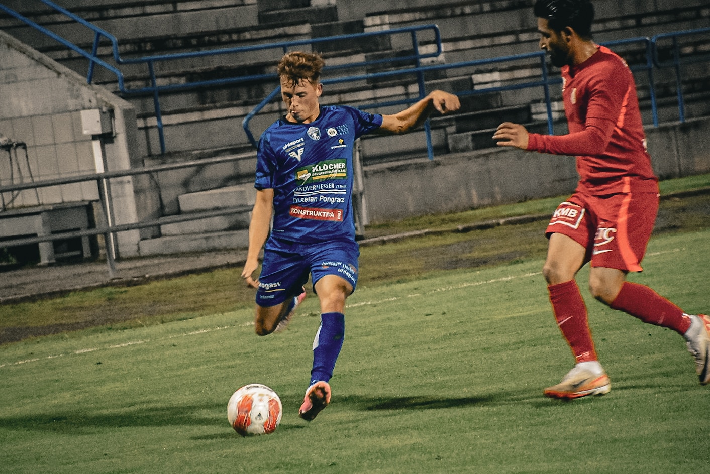 Zwei Fußballspieler kämpfen um den Ball auf dem Spielfeld, einer in blauem Trikot, der andere in rotem Trikot.