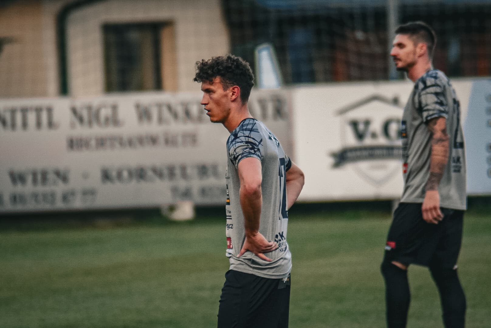 Fußballspieler in grauem Trikot stehen nachdenklich auf dem Spielfeld, Werbetafeln im Hintergrund sichtbar.