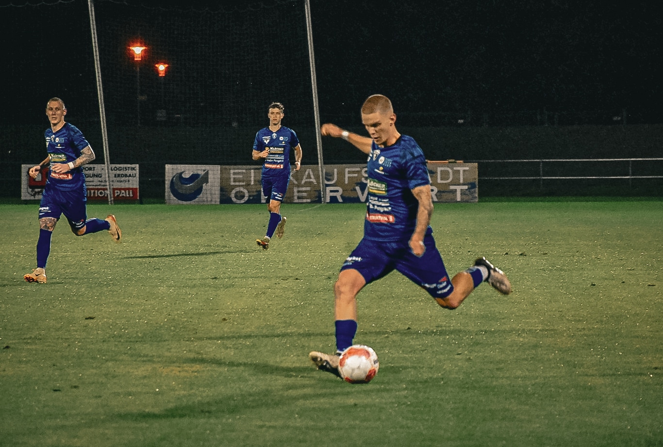 Fußballspieler in blauen Trikots auf dem Spielfeld bei Nacht, einer schießt den Ball.