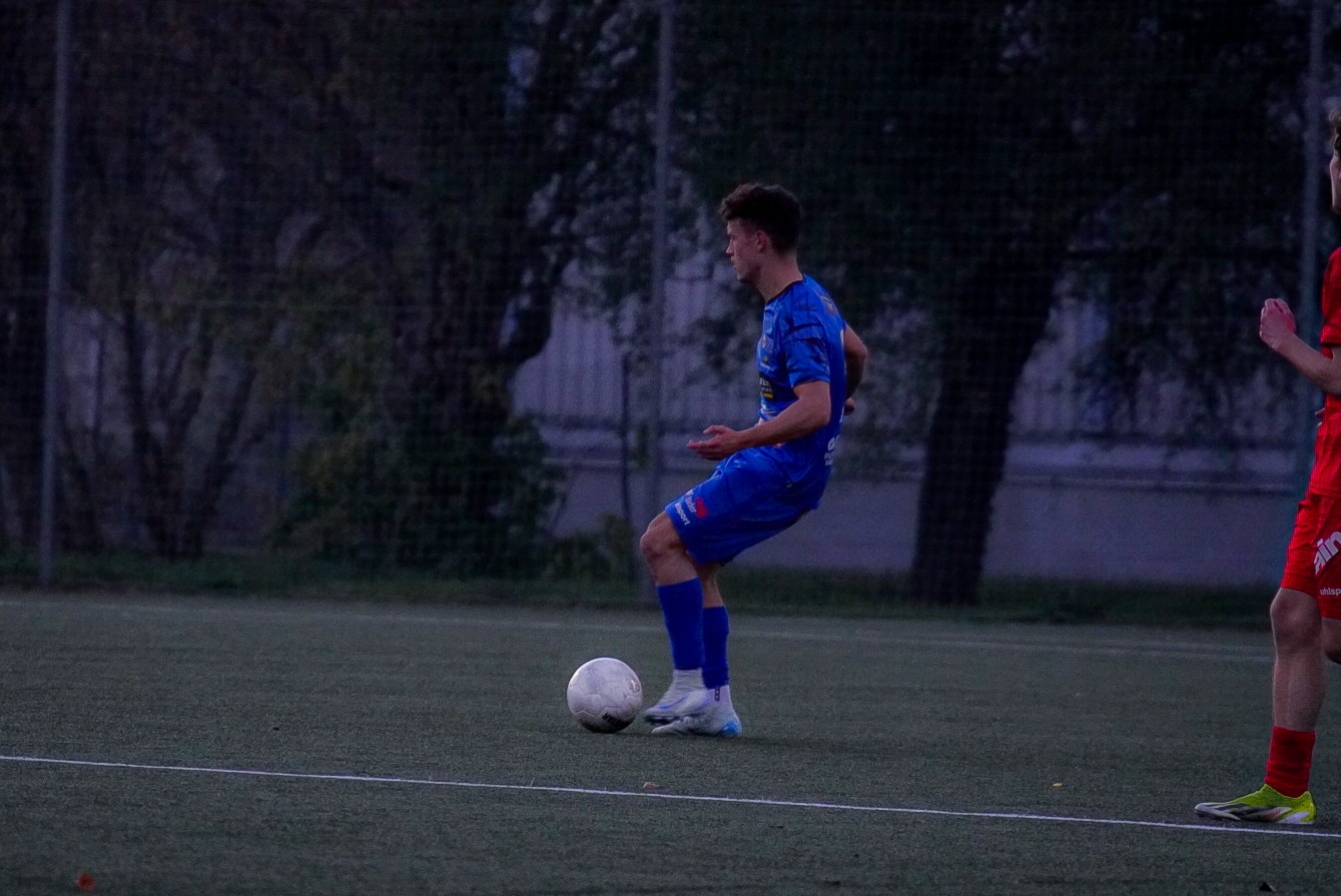 Fußballspieler in blauem Trikot kontrolliert den Ball bei einem Abendspiel auf dem Rasenplatz.