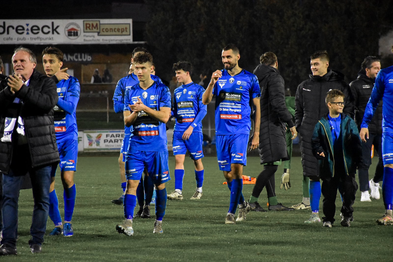 Fußballteam in blauen Trikots feiert auf dem Spielfeld bei Nacht. Spieler klatschen und lachen.