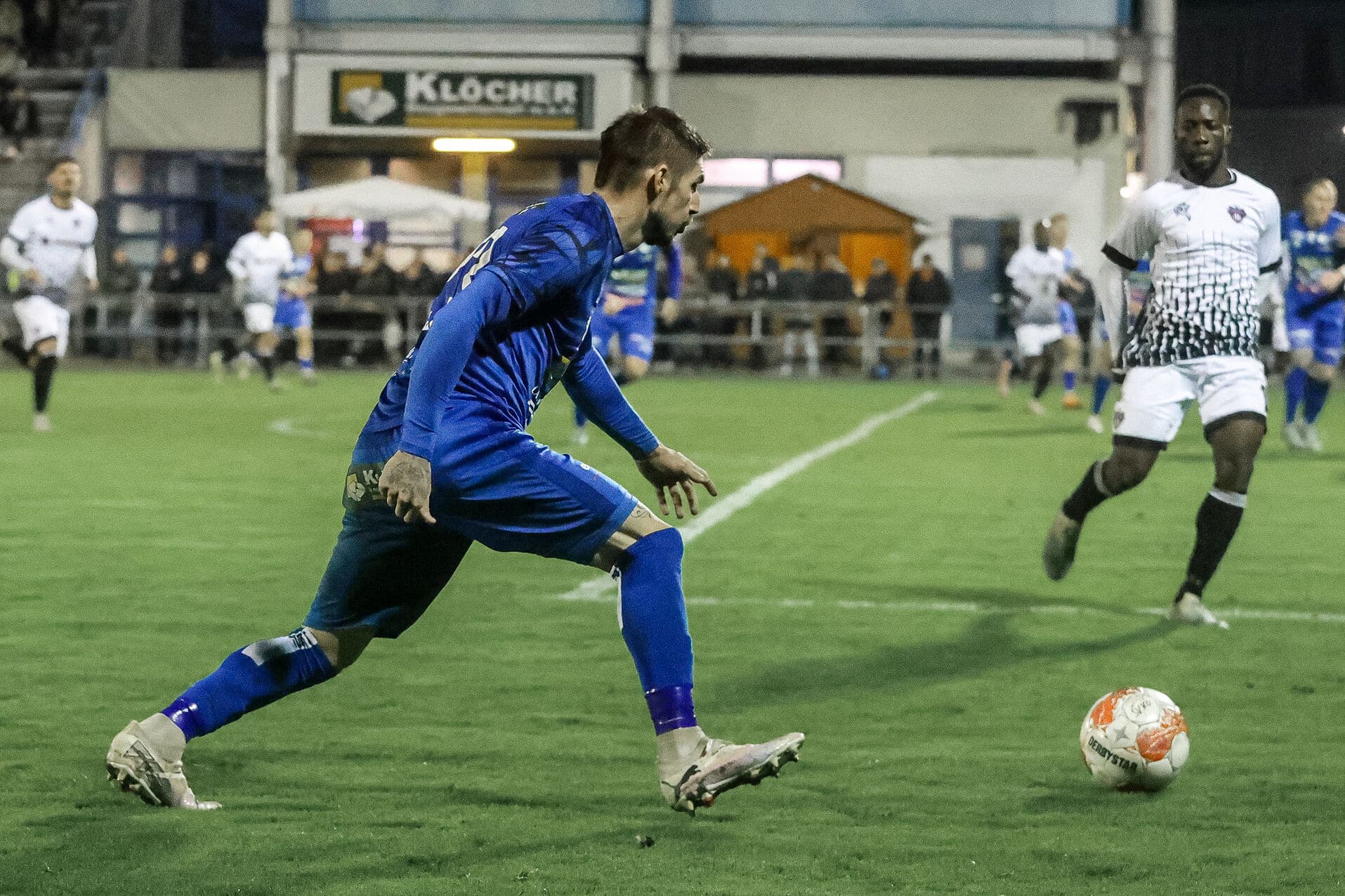 Fußballspieler in blauem Trikot rennt mit Ball über das Feld, während Gegner in schwarz-weißem Trikot hinterherläuft.