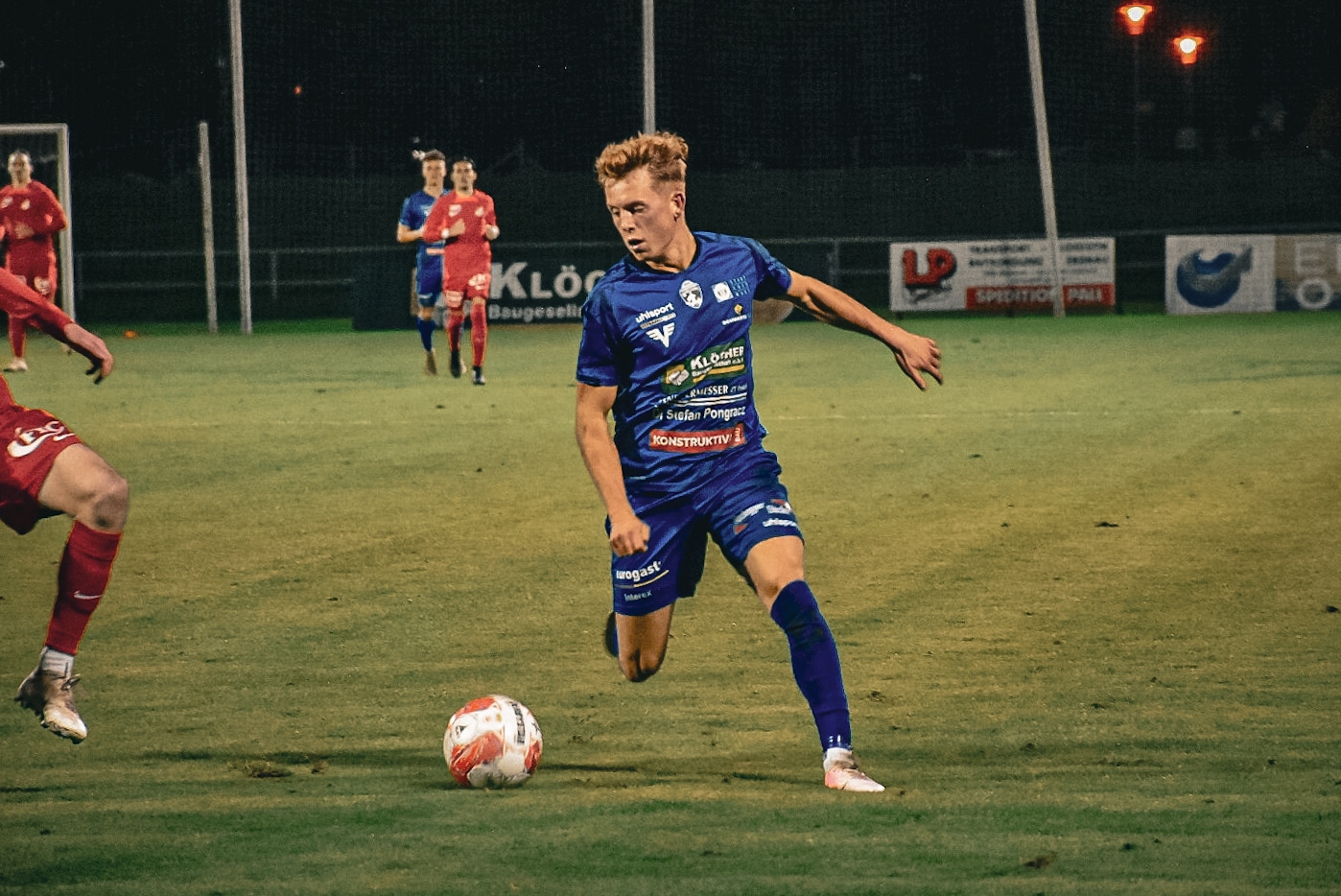 Junger Fußballspieler in blauem Trikot dribbelt den Ball während eines Abendspiels auf einem beleuchteten Fußballfeld.